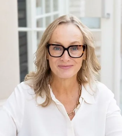 A middle-aged woman with blonde hair, glasses, and hoop earrings, sitting indoors near windows with a white background.