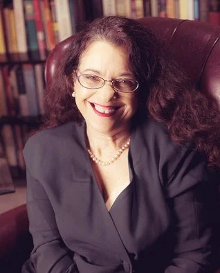 A woman with dark, curly hair and glasses smiling, sitting in a leather chair with bookshelves in the background.