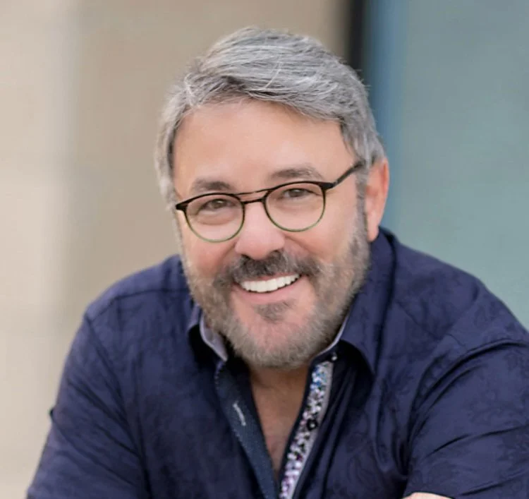 A middle-aged man with gray hair, glasses, and a beard smiling in a close-up portrait.