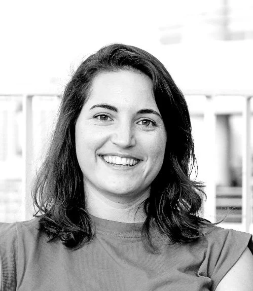 Black and white photo of a woman with shoulder-length dark hair, smiling, wearing a crew neck shirt, with a blurred building background.