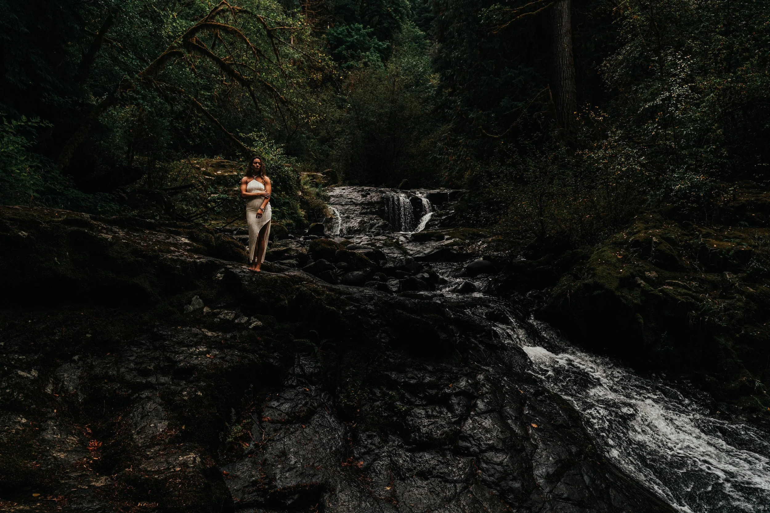 A woman in a white dress standing on rocks beside a waterfall in a lush, green forest.