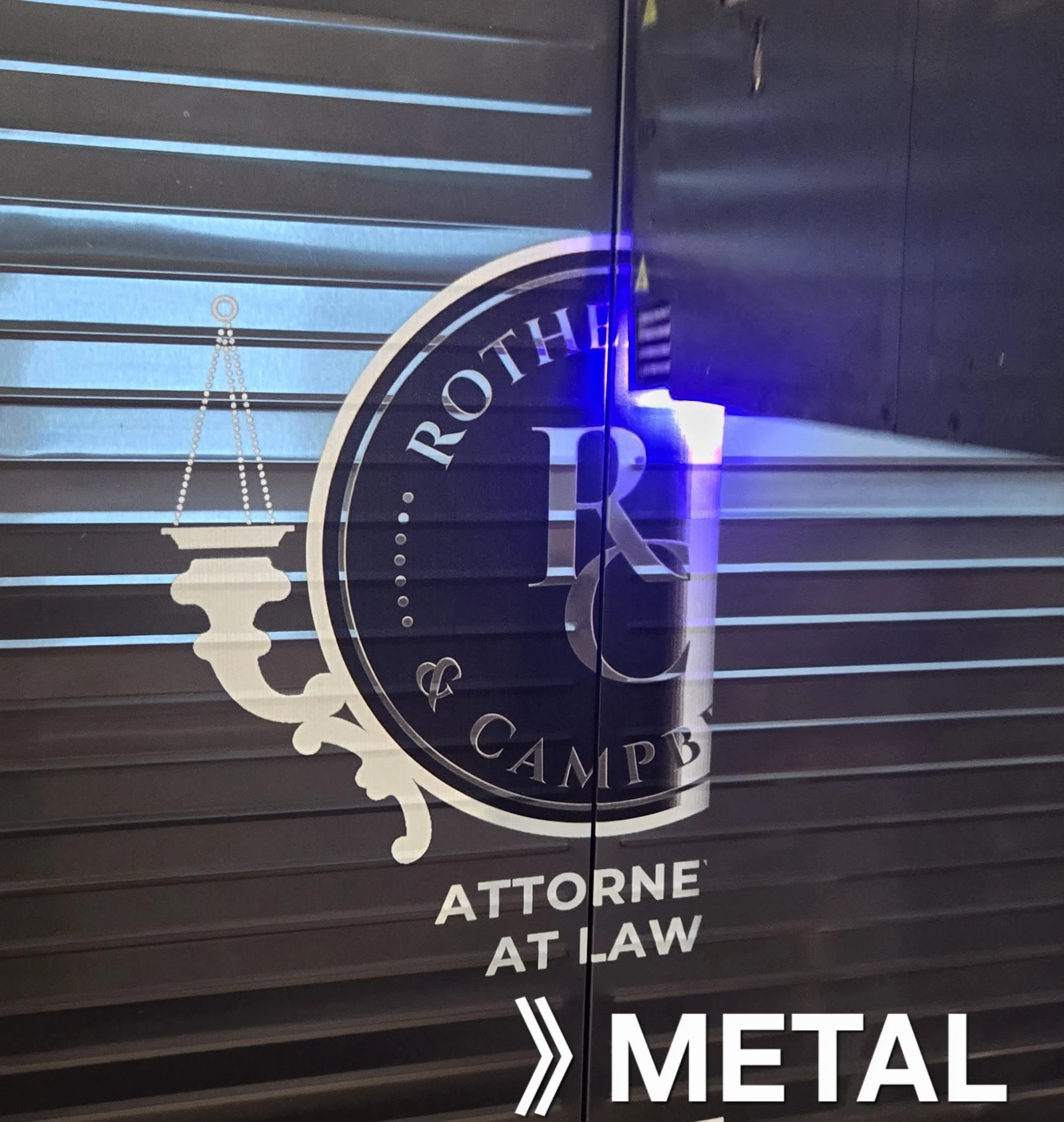 Sign with text 'The Rother Camp' featuring a colonnade, with additional text 'ATTORNE AT LAW' below, displayed on a metal surface.