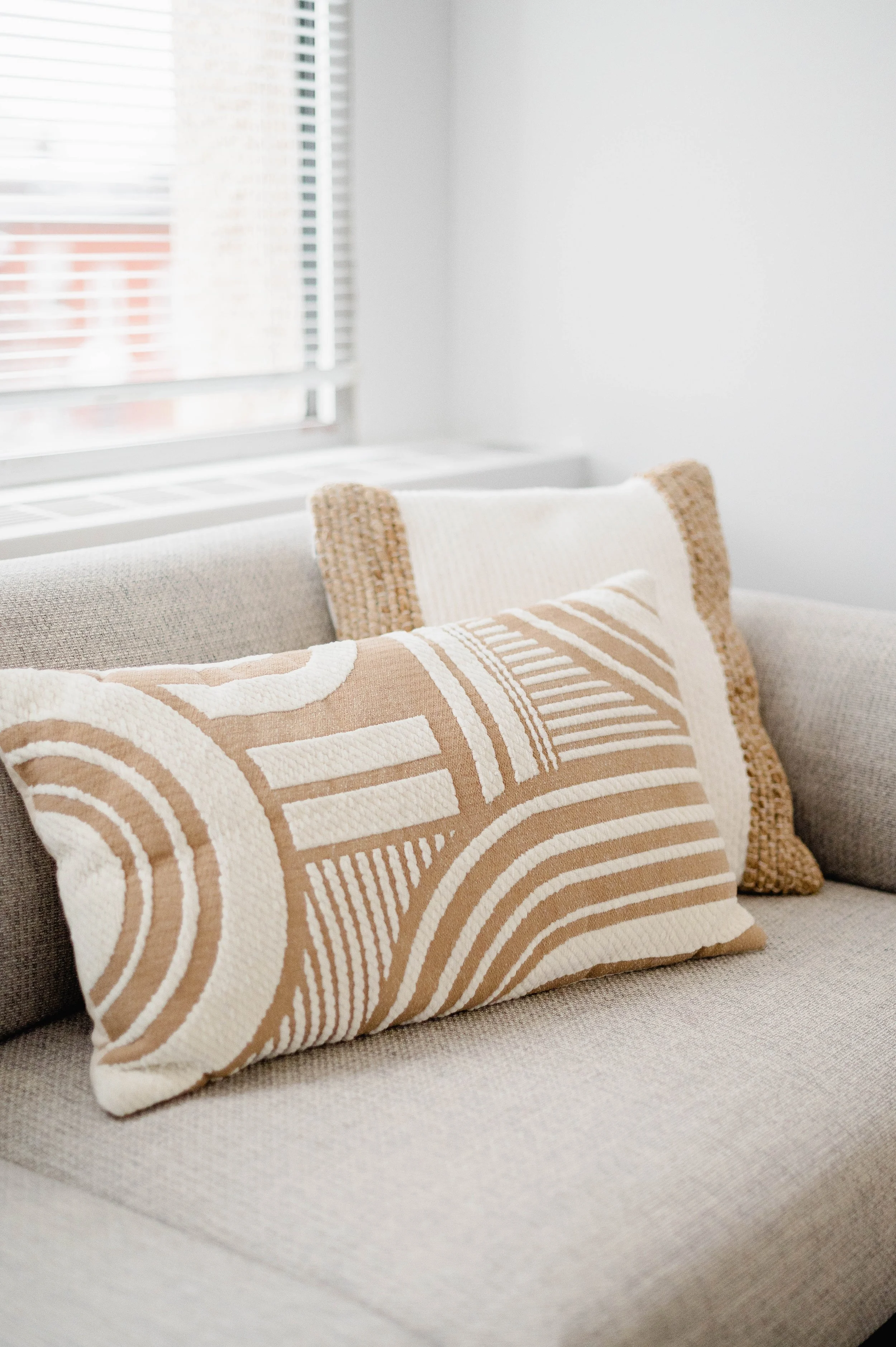 Close-up of a beige and white decorative pillow on a light gray sofa near a window.