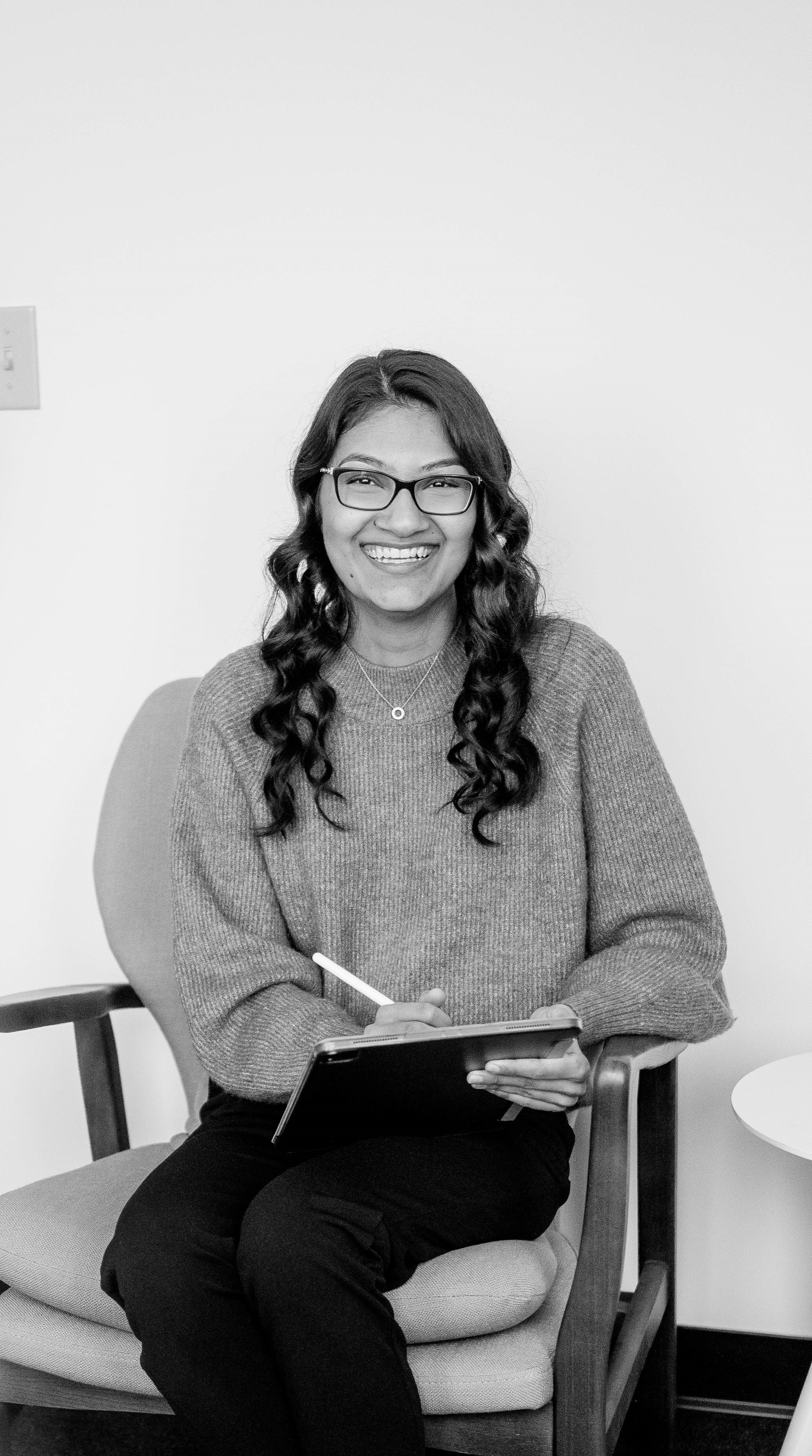 A kind woman smiling while sitting in a chair, holding a tablet in a therapy room.