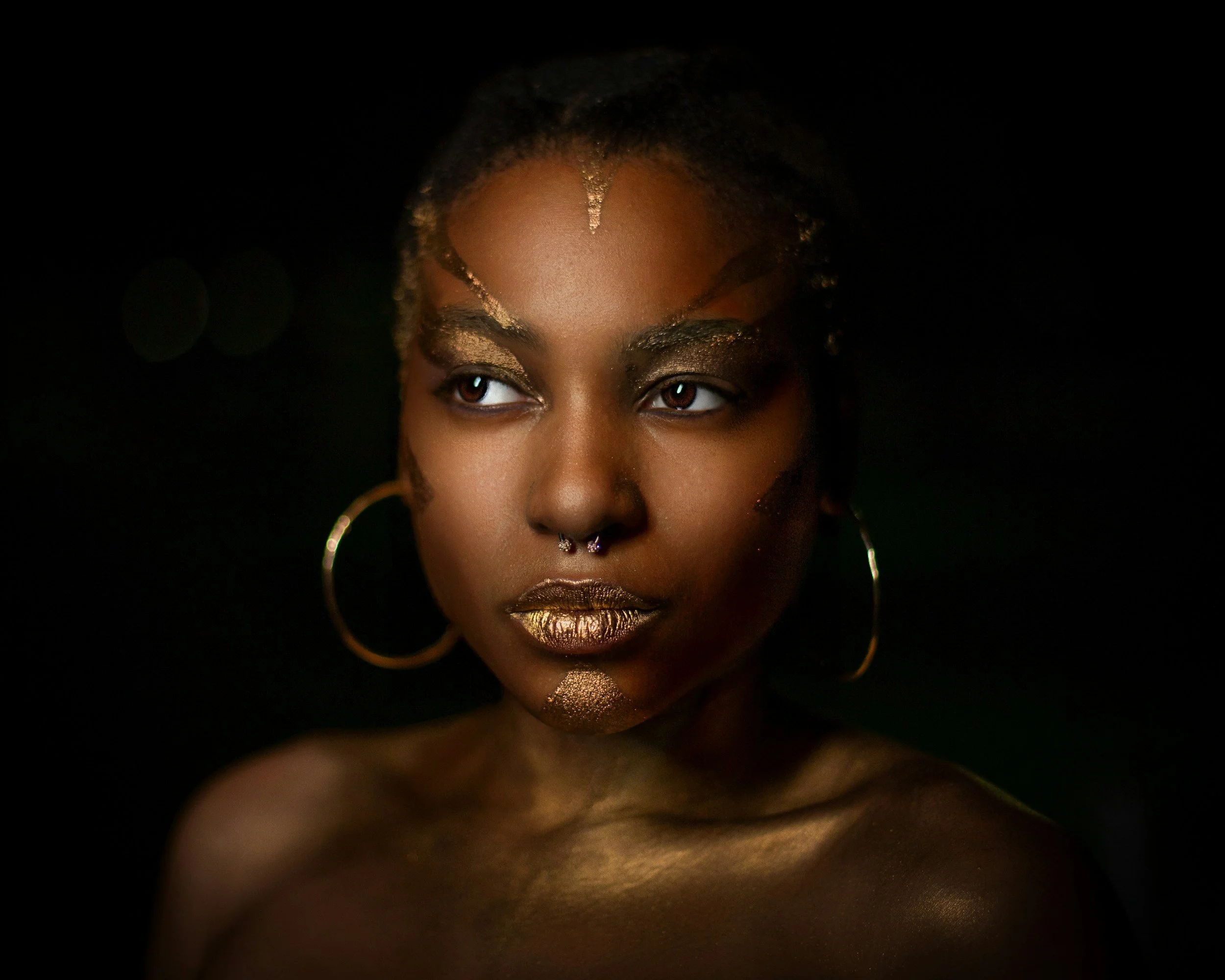 Close-up of a woman with dark skin and gold makeup, gold lipstick, gold glitter on her face, large gold hoop earrings, and gold makeup accents on her eyebrows and face, against a black background.