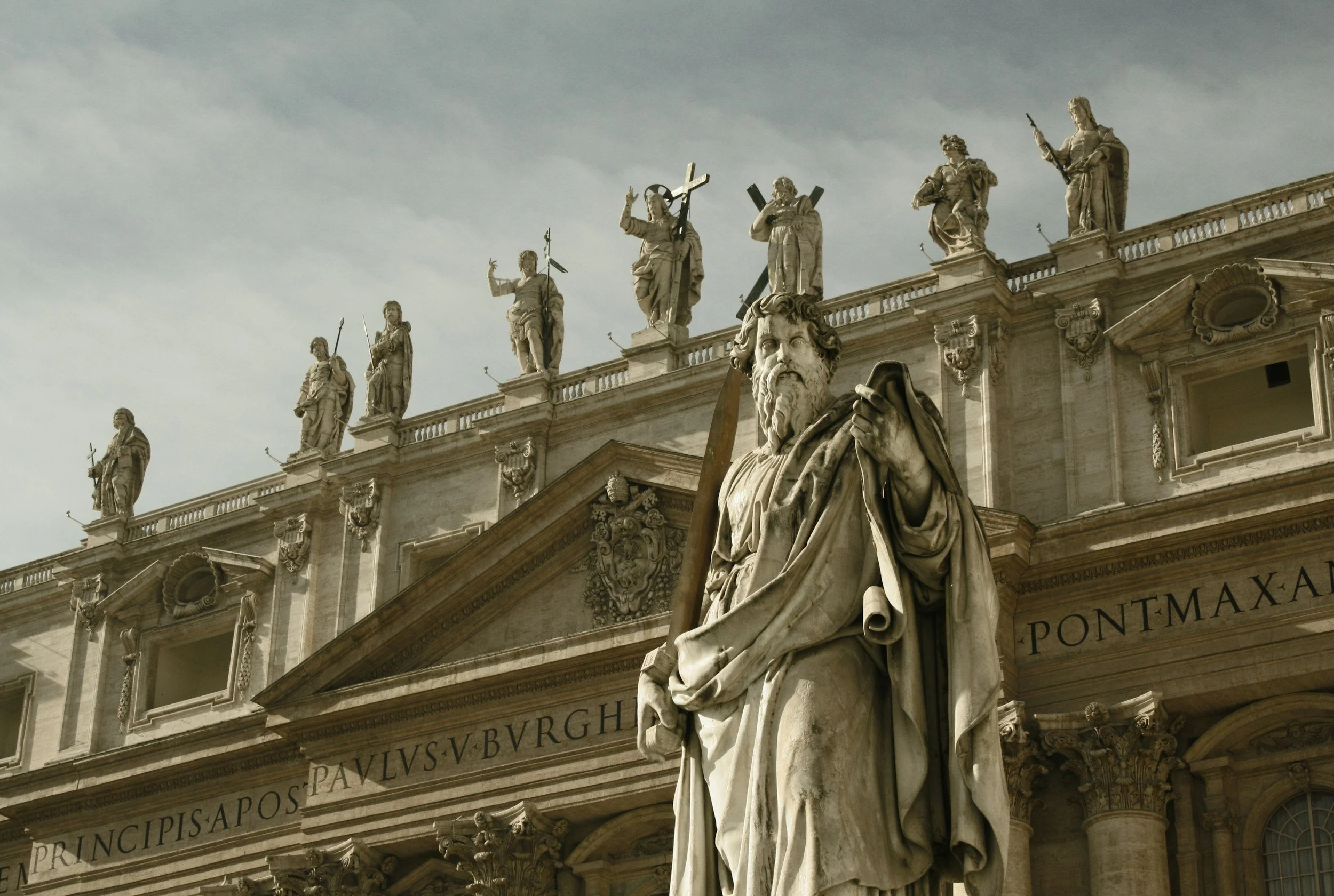 Stone statue of a bearded man holding a staff and a scroll, in front of a historic building with sculptures of saints or religious figures on the roofline.
