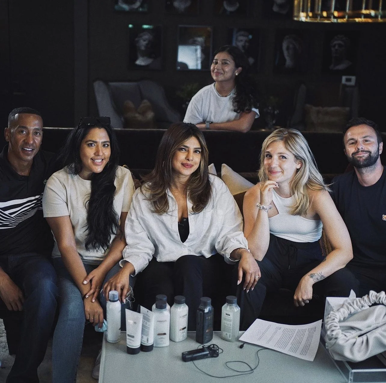 Group of six diverse adults sitting on a couch and a woman standing behind them in a dimly lit room, with bottles and papers on the table in front including Priyanka Chopra-Jonas
