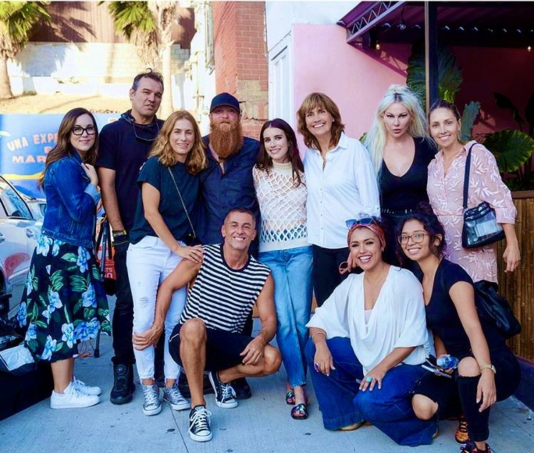 Group of ten diverse friends posing outdoors on a city sidewalk, smiling at the camera including celebrity Emma Roberts.