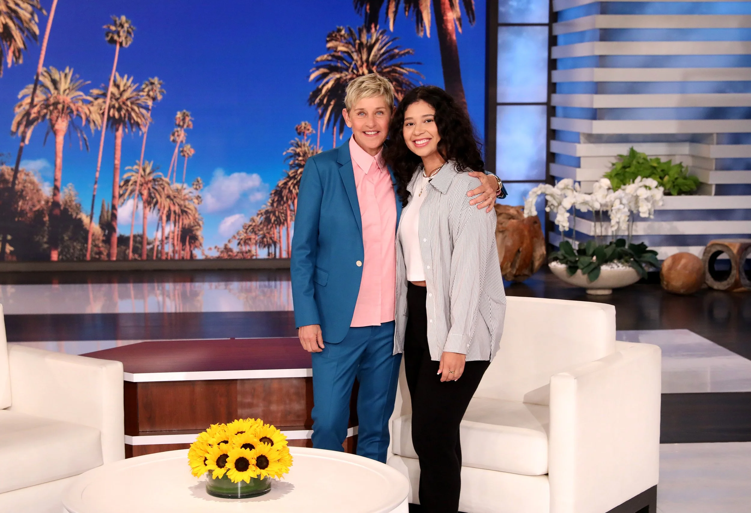 Two women standing together on a TV show's set, smiling for the camera, with palm trees and a blue sky background, and sunflowers on a table in the foreground including Ellen DeGeneres.