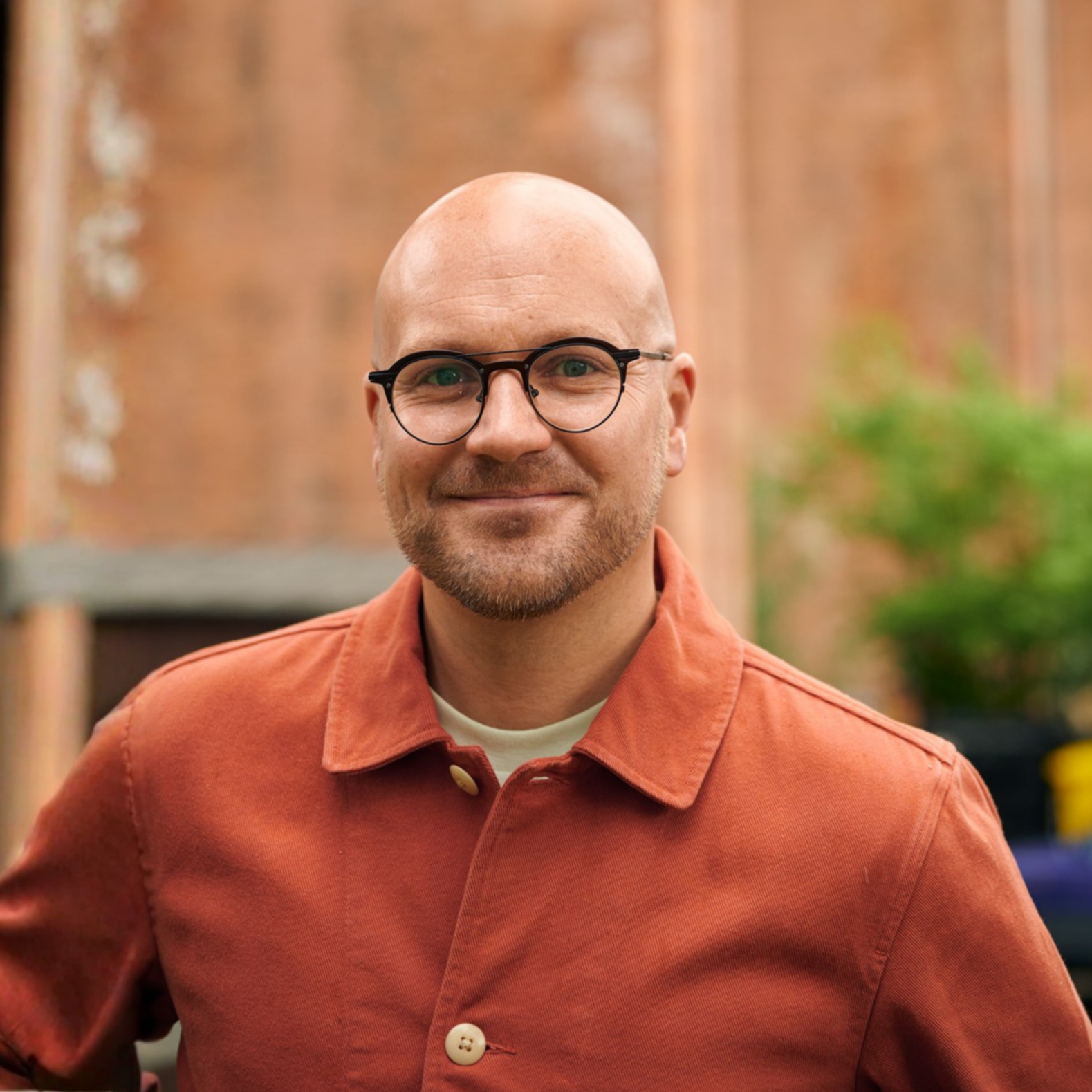Smiling man with glasses wearing a rust-coloured jacket, standing outdoors against a blurred brick background.