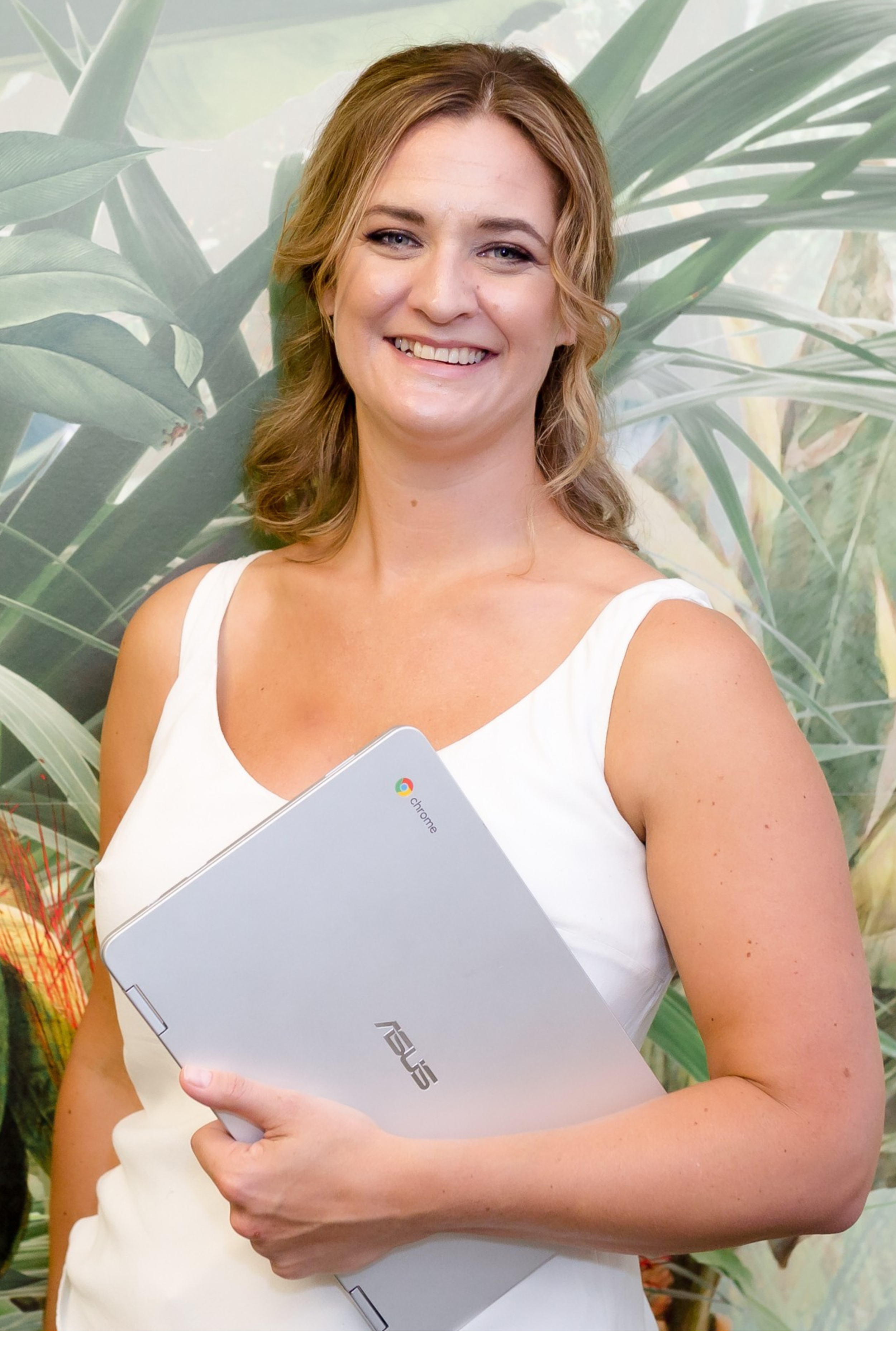 Smiling woman in a white sleeveless top holding a silver laptop, standing before a leafy green backdrop.