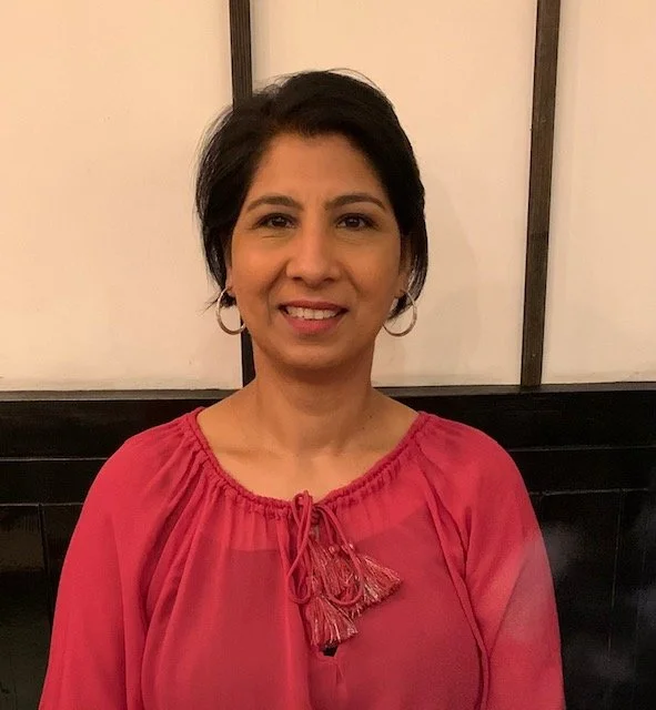 Smiling woman with short dark hair wearing a pink blouse, standing indoors against a light wall.