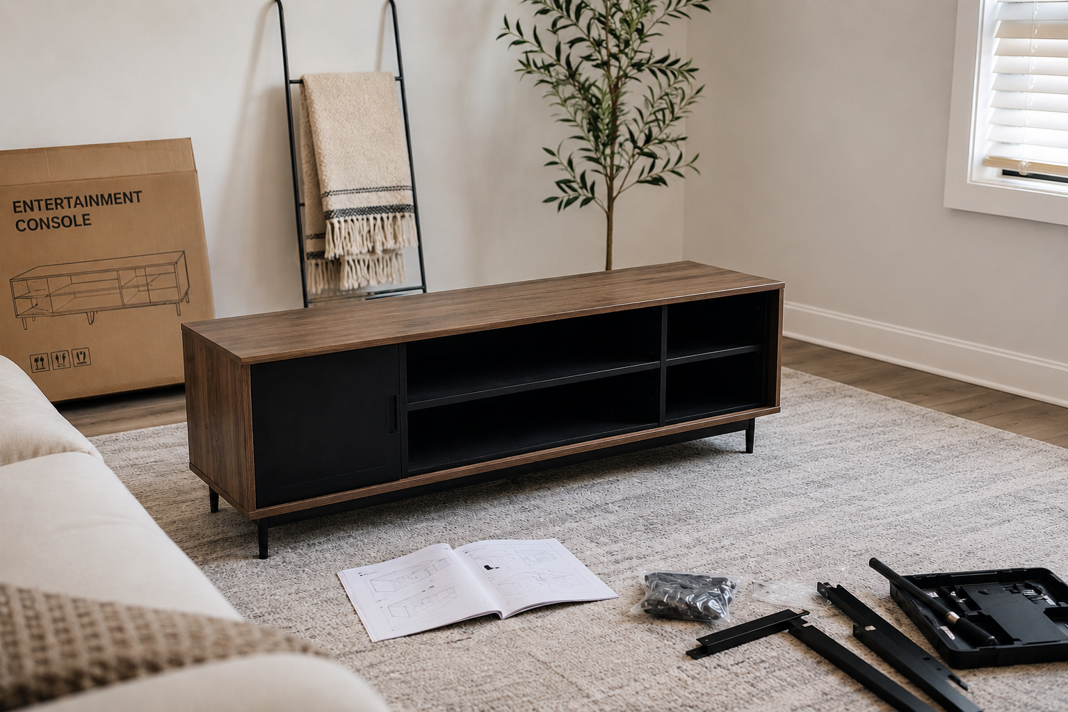 Living room with a wooden TV stand, open instruction manual, black hardware pieces, and tools on a light-colored rug. A cardboard box labeled 'Entertainment Console' is in the background along with a tall plant, a blanket on a black towel rack, and a window with white blinds.