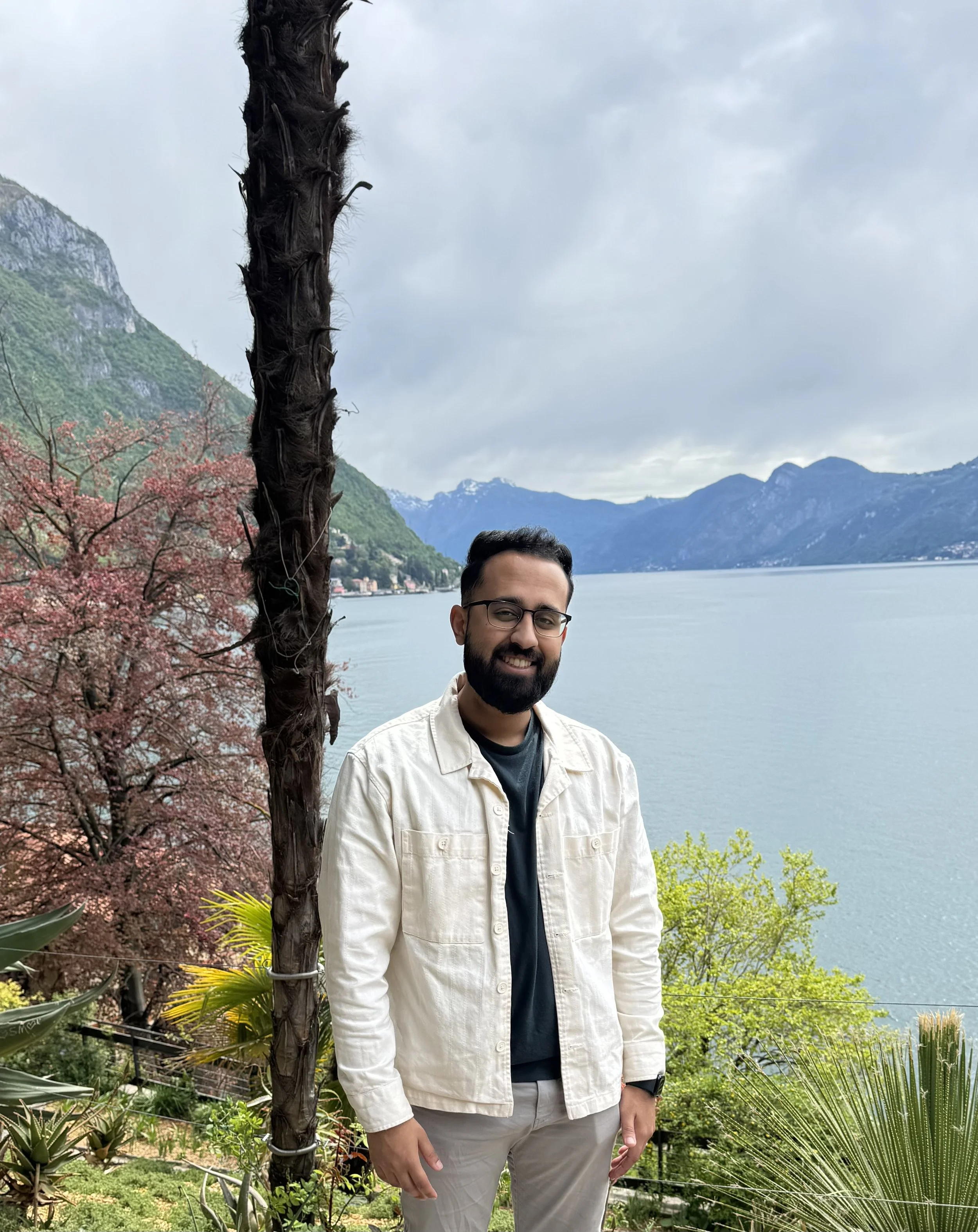 A man with glasses and a beard standing outdoors near a tree with mountains and water in the background on a cloudy day.
