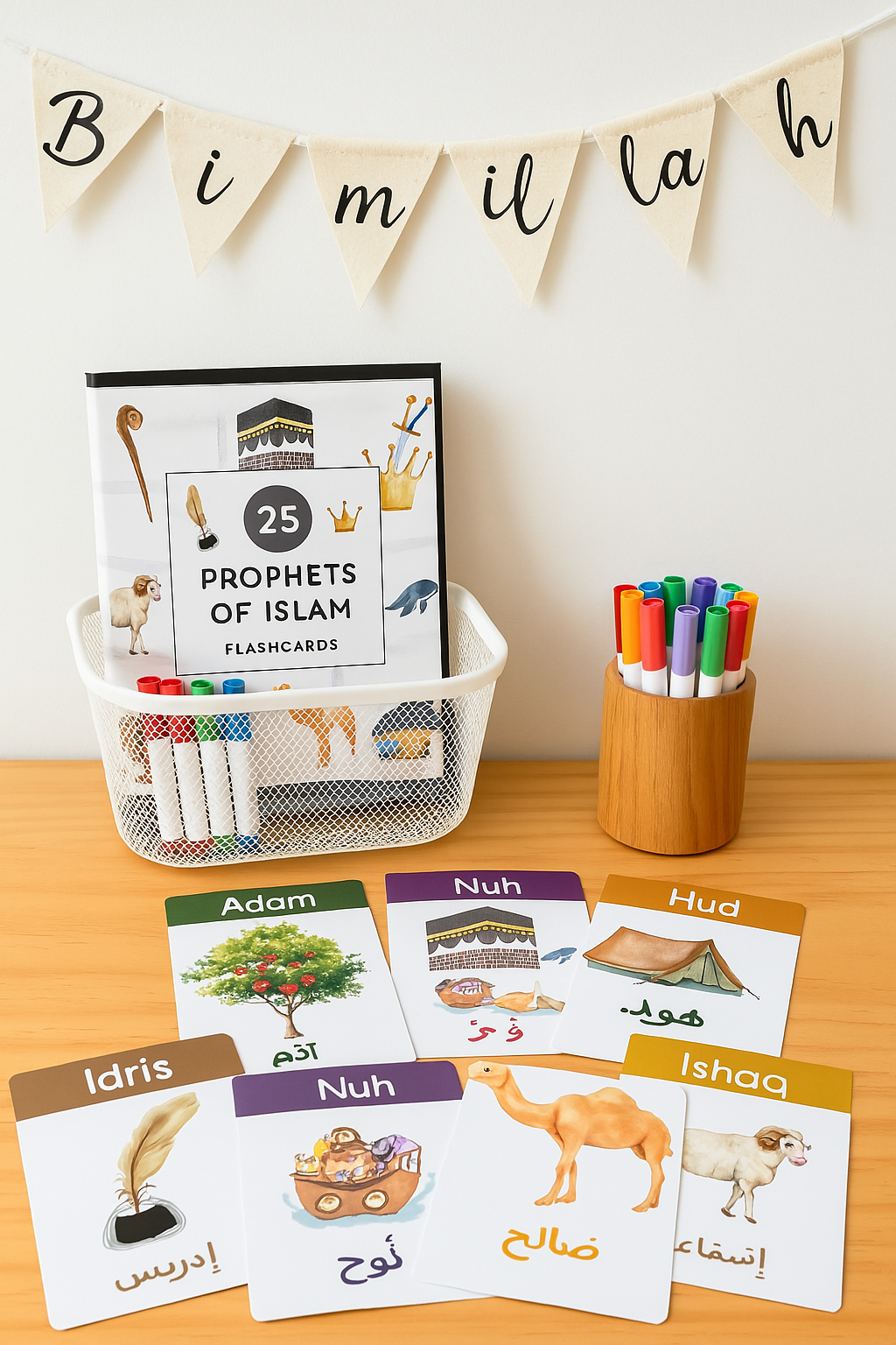 Educational display featuring flashcards about the prophets of Islam. The scene includes a banner that says 'Bimillah', a book titled '25 Prophets of Islam Flashcards', and colorful markers in a wooden container. Several flashcards with images and names of prophets like Adam, Nuh, Hud, Idris, and Ishaq are spread out on a wooden surface.