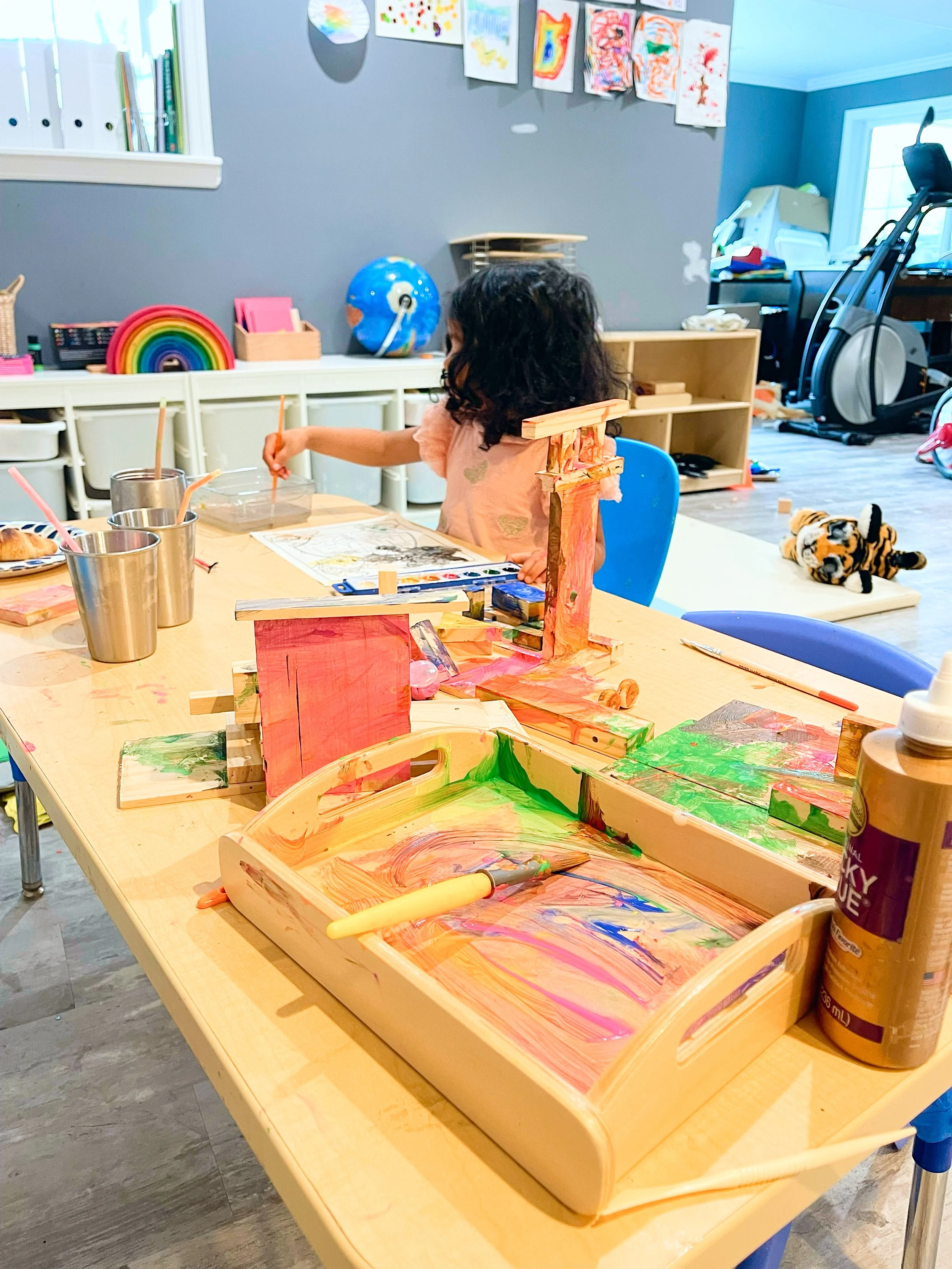 A young girl at a table engaged in arts and crafts with wooden blocks and paint. The table is covered with colorful paints, brushes, and wooden crafts. In the background, there is a shelf with art supplies, a blue globe, and a plush tiger toy on the floor near a treadmill.