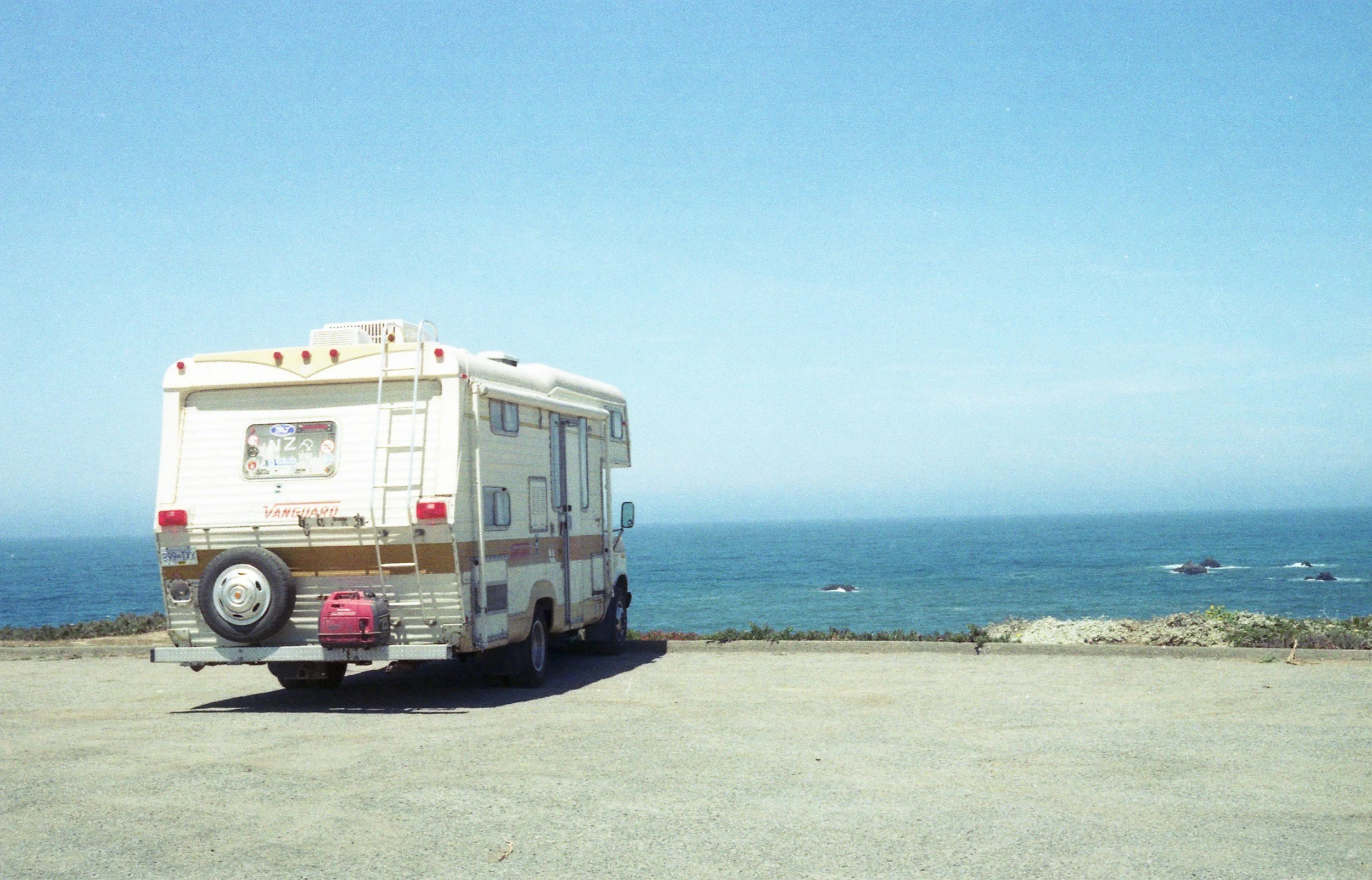 A white camper van parked on a gravel lot overlooking the ocean under a clear blue sky.