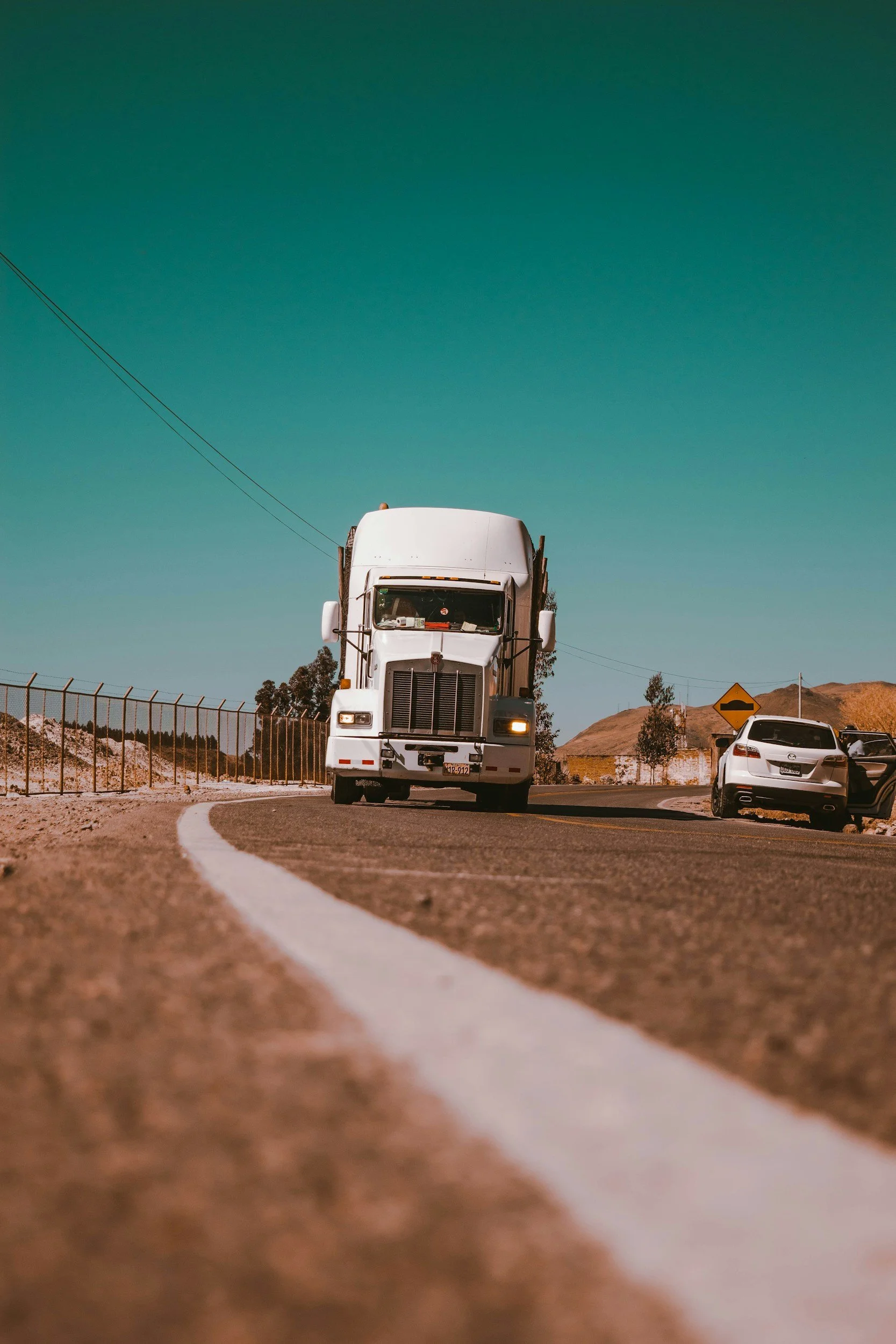 A white semi-truck driving on a curved road with a car stopped on the side, under a clear blue sky and dry landscape.