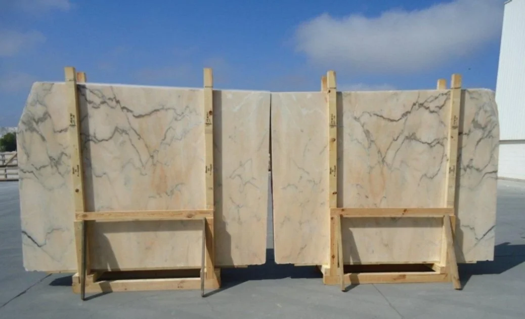 Large marble slab with a light beige color and dark veining, supported by wooden racks outdoors under a blue sky with some clouds.