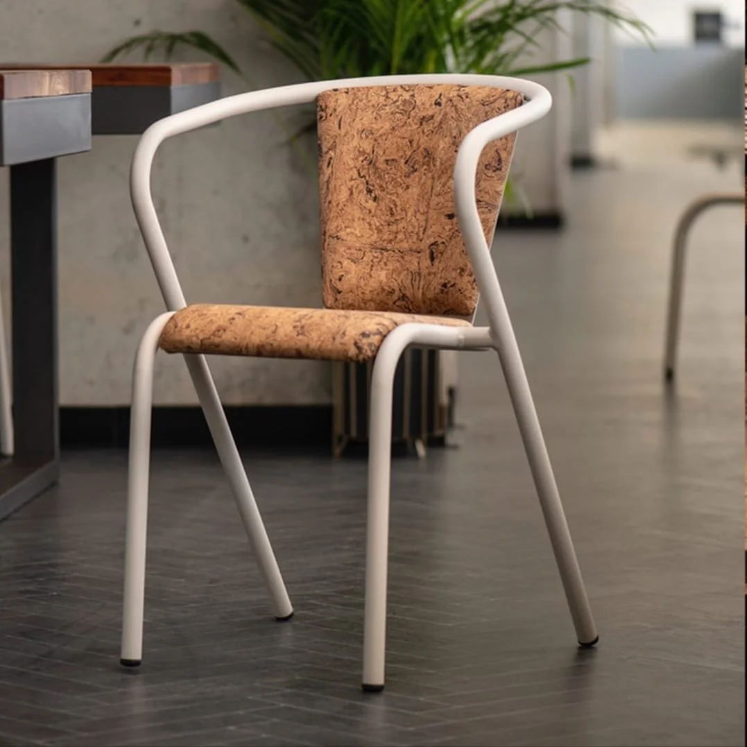 A chair with a white metal frame and a cork seat and backrest, placed on a dark wooden floor in a modern interior with plants and furniture.