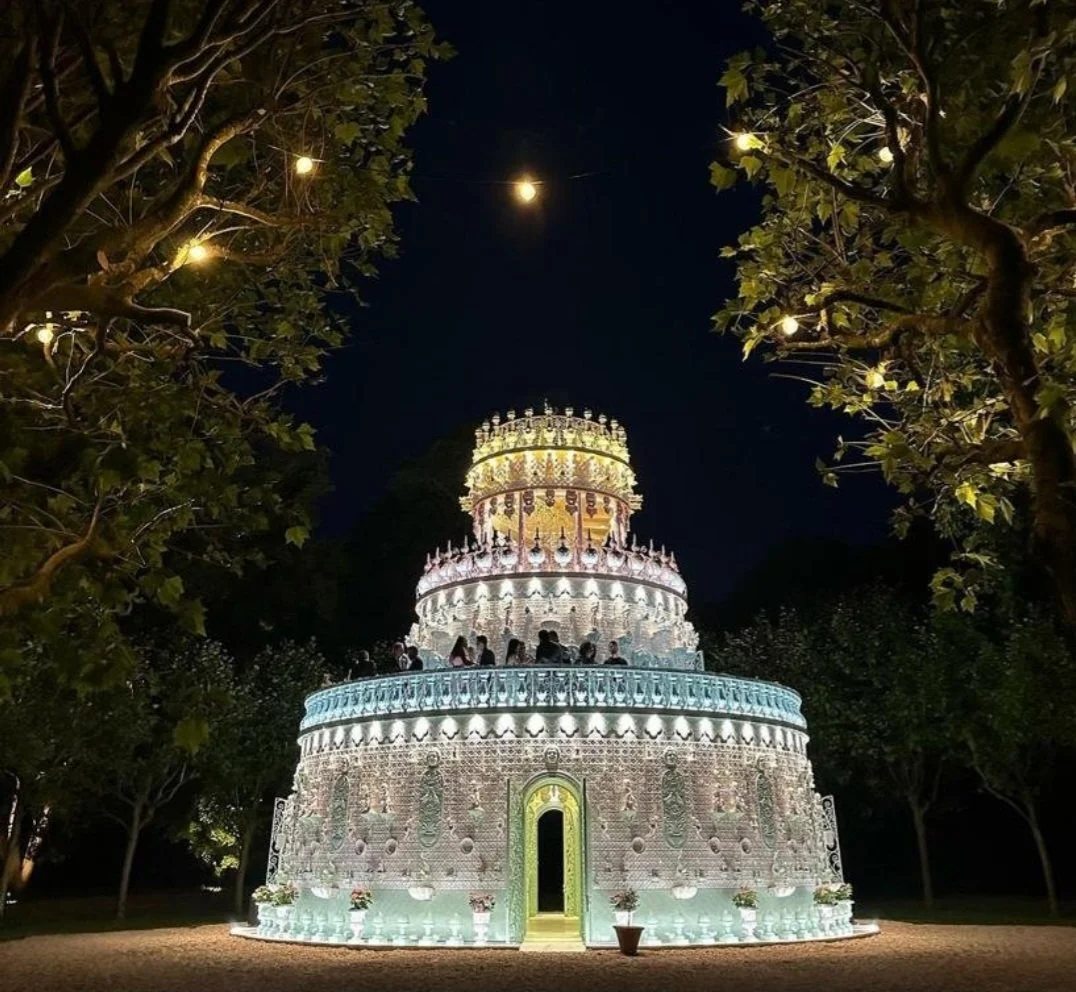 A brightly lit, multi-tiered decorative structure resembling a cake at night, surrounded by trees and outdoor string lights.