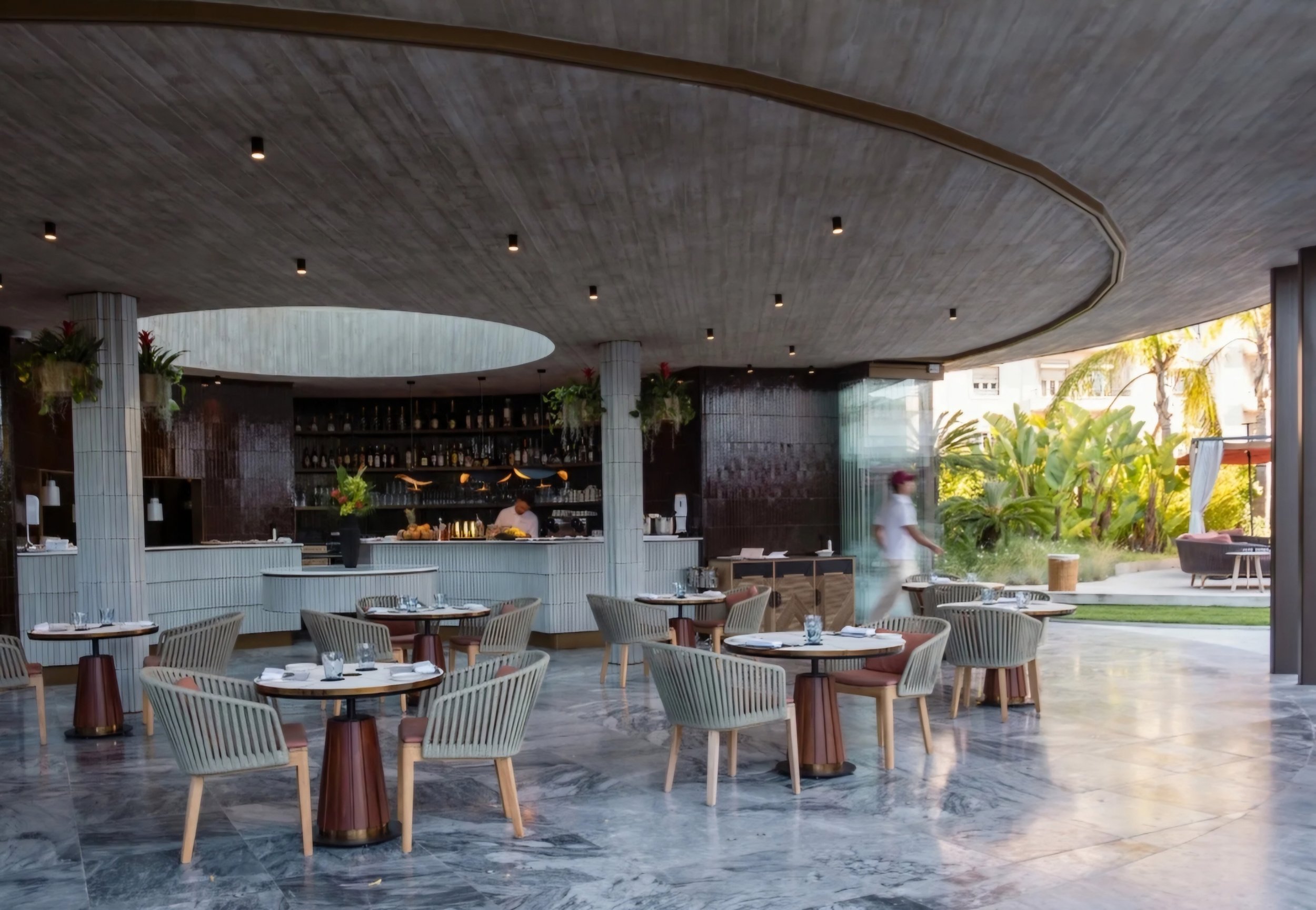 Modern restaurant interior with round tables, chairs, plants, and a bar area with bottles on shelves.