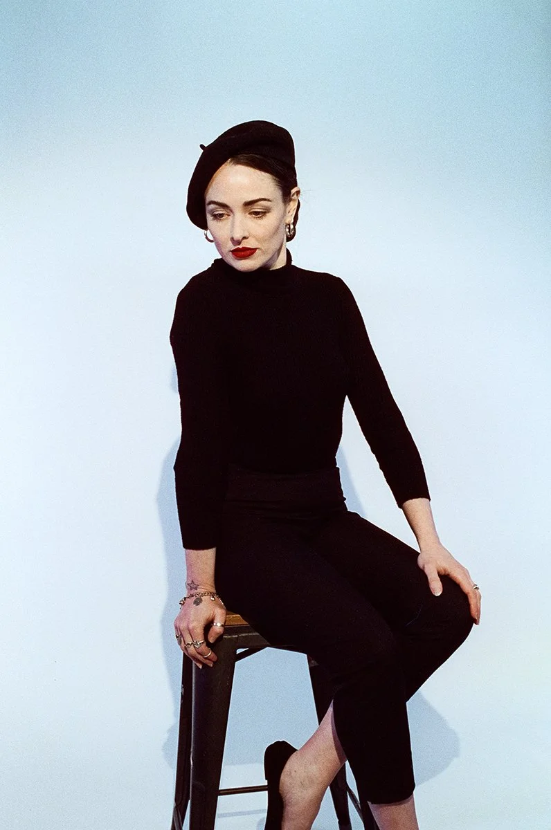 A woman dressed in black, wearing a beret, sitting on a stool against a plain white background.