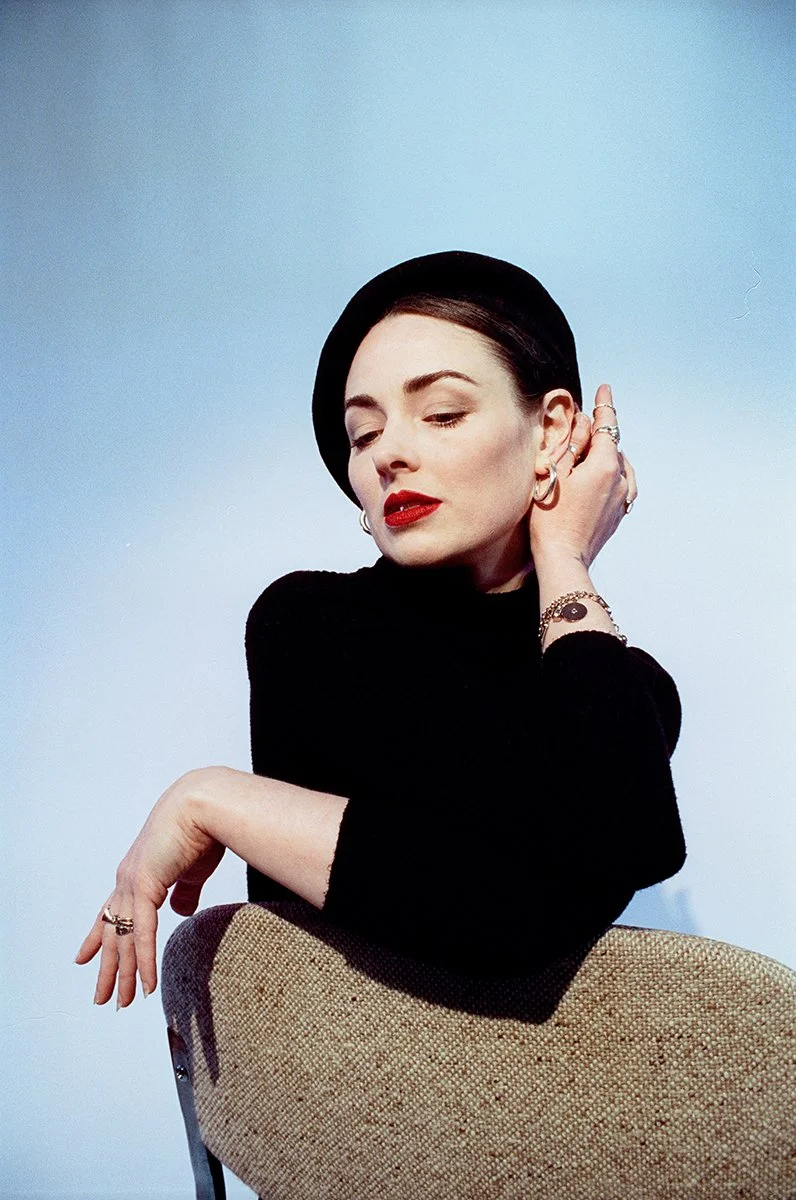A woman with dark hair, red lipstick, wearing a black beret and black top, sitting on a beige chair against a plain light blue background.