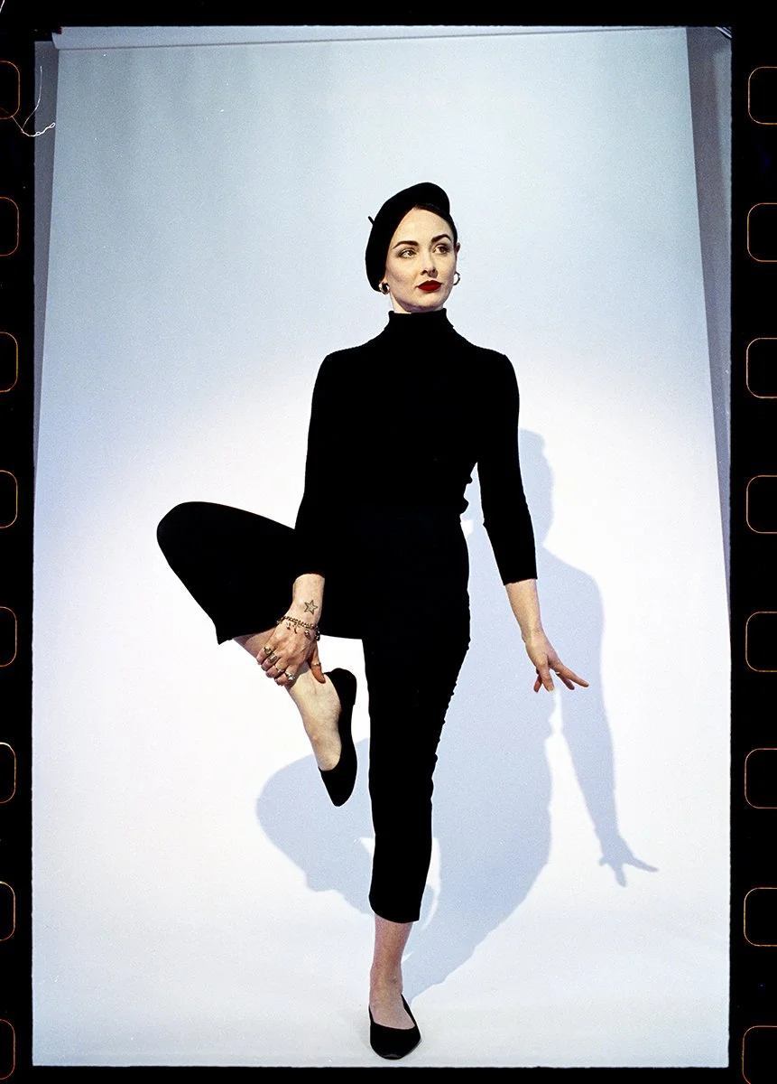 A woman with fair skin, dark eyebrows, red lipstick, wearing a black turtleneck and pants, black beret, black flats, and jewelry, standing against a white background in a yoga pose with one leg bent and her hand holding her foot, casting a shadow beh