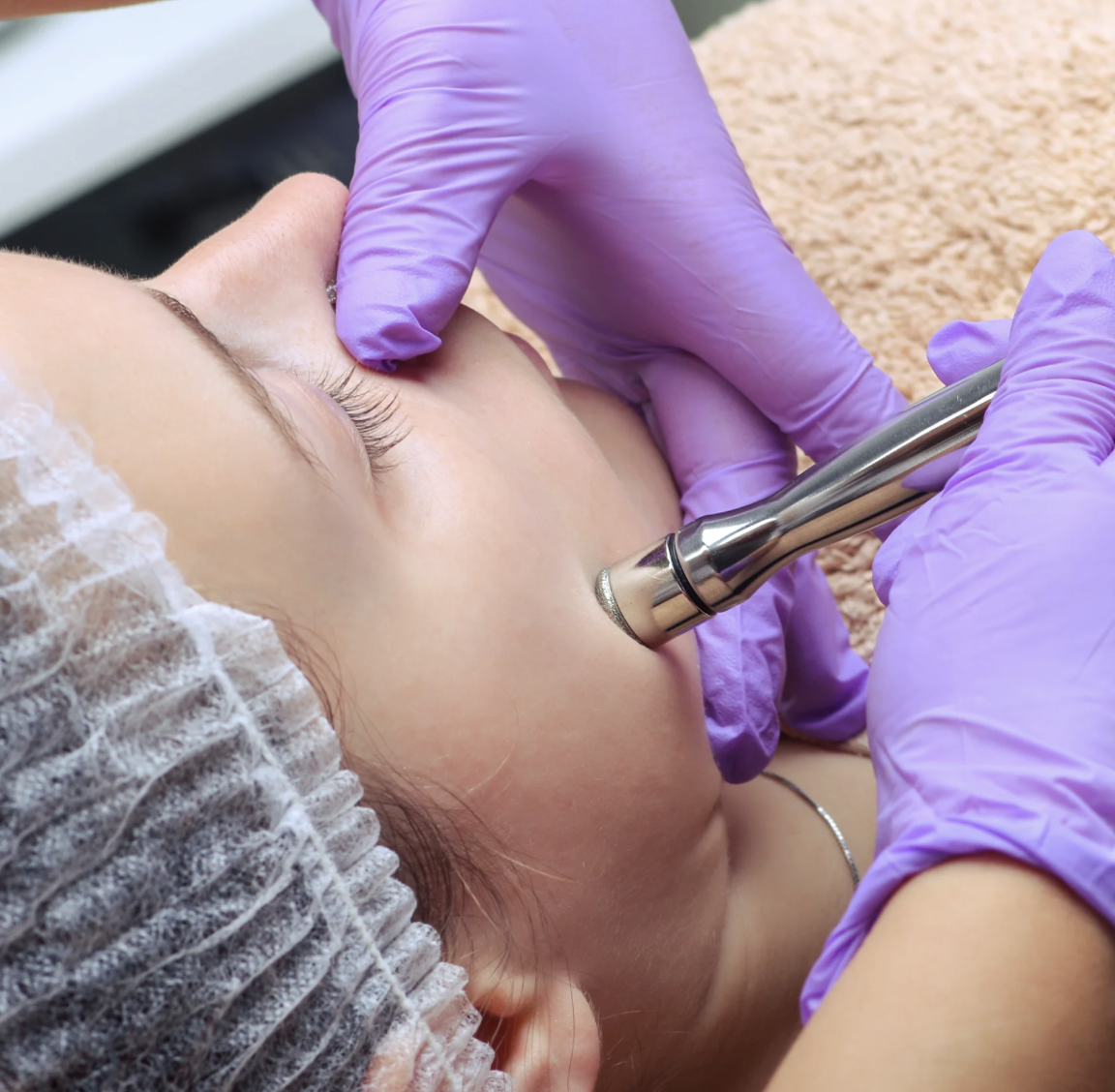 A person receiving a facial treatment with a skincare device in a spa setting, wearing a hair cap and purple gloves.