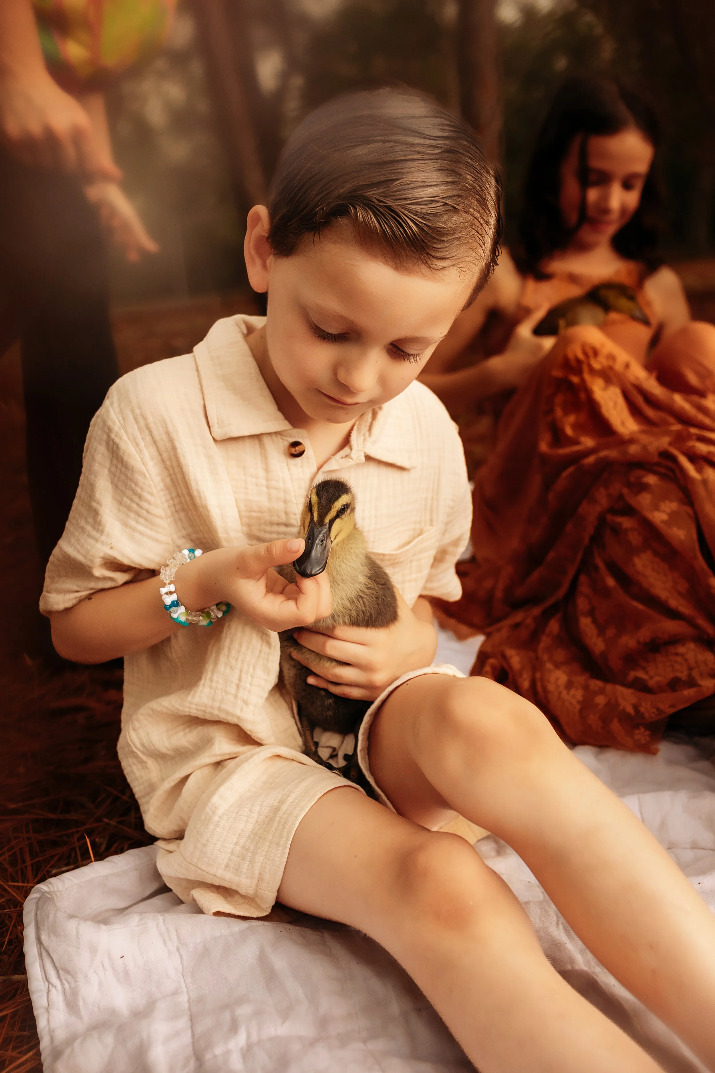 A young boy with wet hair holding a duckling, sitting on a white cloth in a natural outdoor setting with trees in the background. A girl in a patterned dress is in the background looking at the duckling.