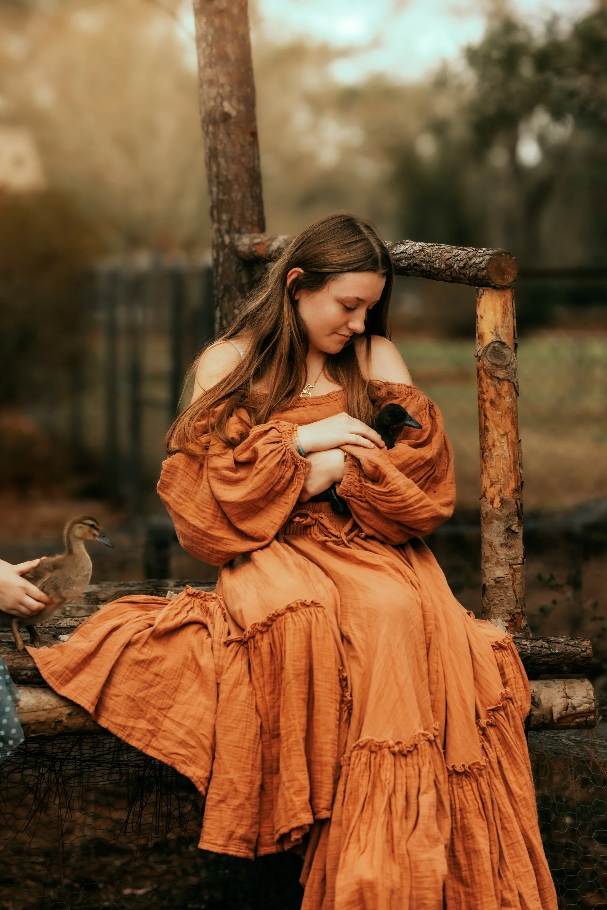 A woman with long brown hair, wearing an orange dress, is sitting on a log outdoors holding a black duck close to her chest. Another duck is standing nearby, being held by someone out of frame. The background features trees and a blurred, natural set