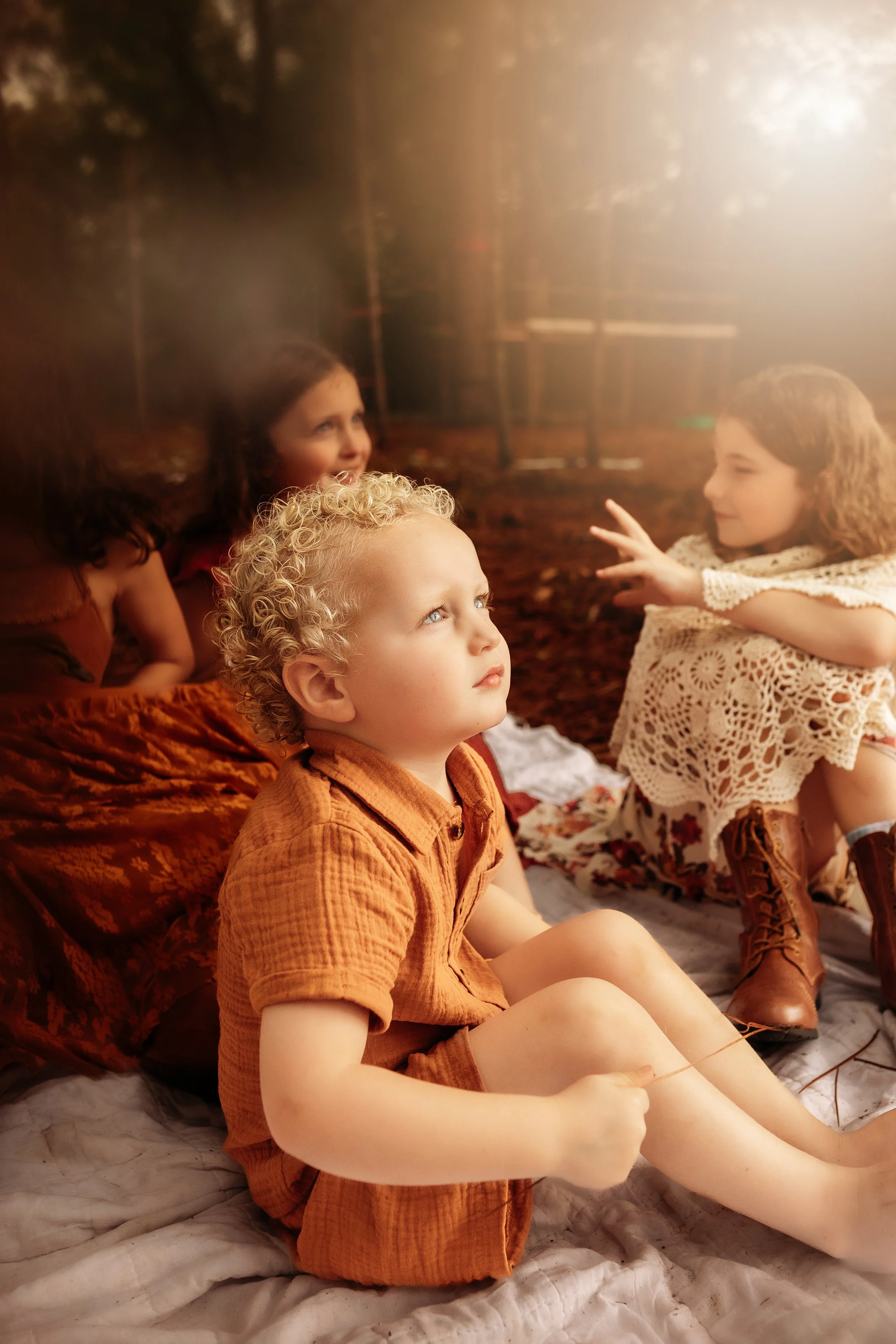 Three children with curly hair sitting on a soft surface, with a girl in a cream lace dress reaching out to a girl with dark hair, and a boy in an orange shirt looking up.