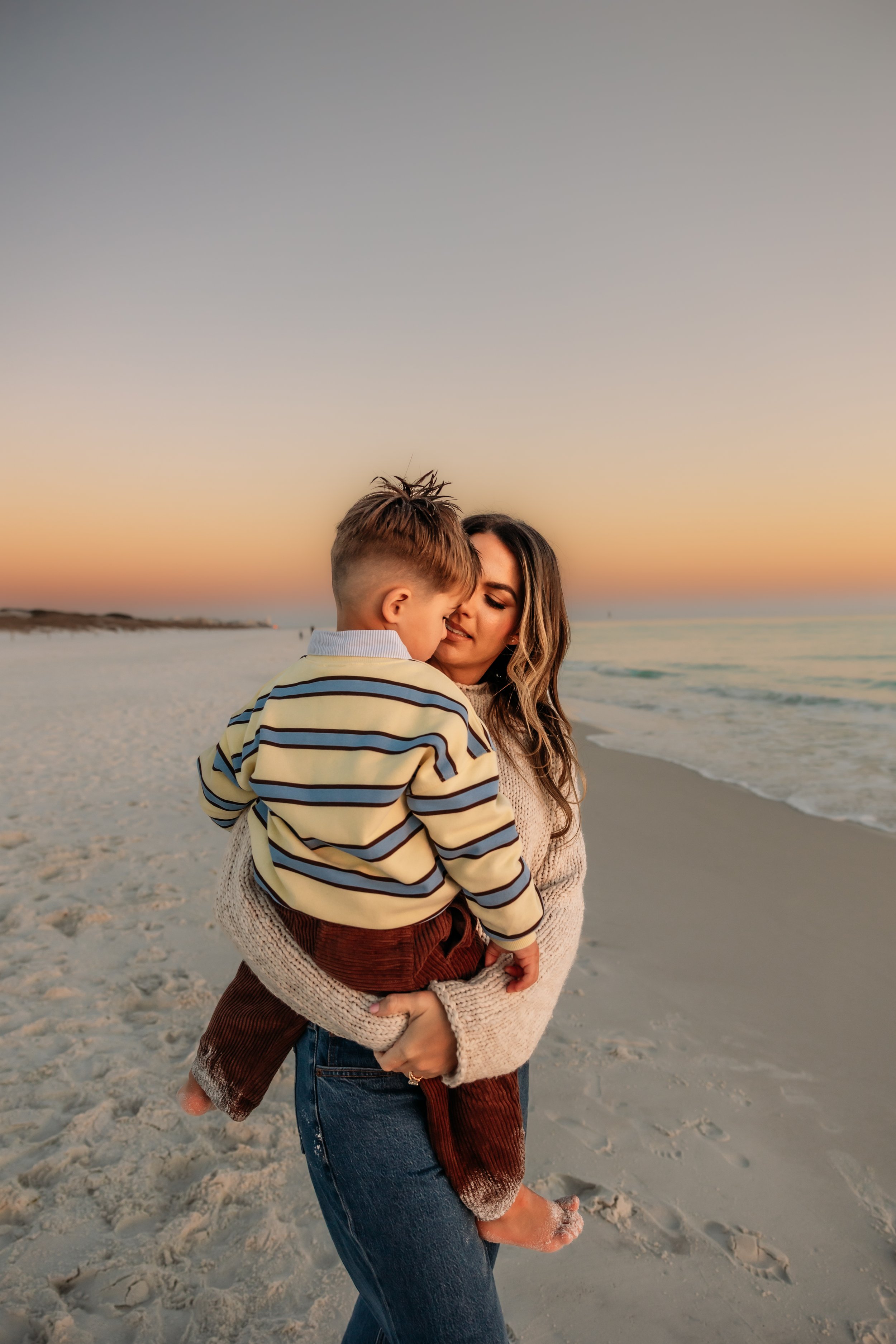 A woman holding a young boy at the beach during sunset, with the ocean and sandy shore in the background.