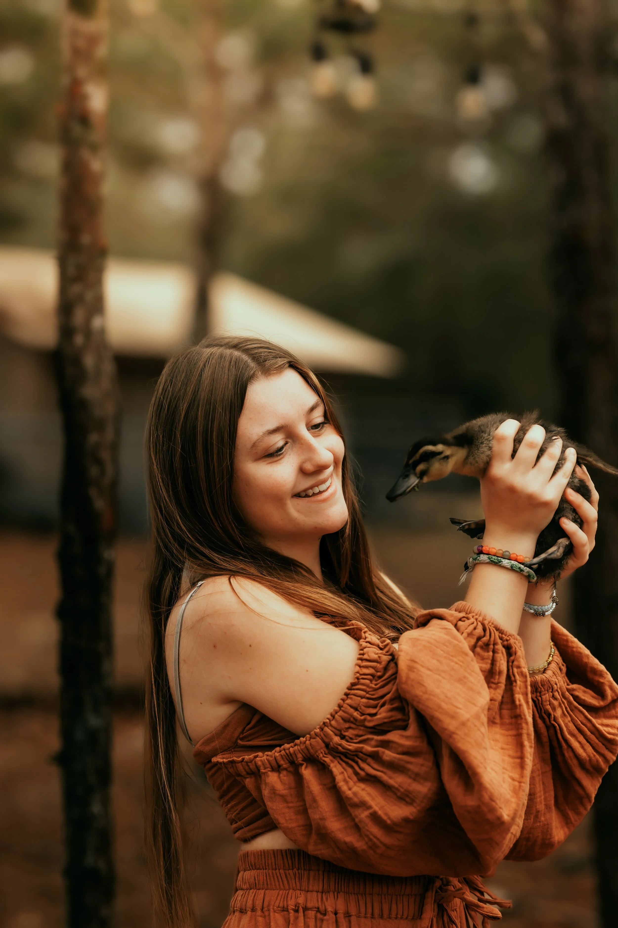 A young woman with long brown hair smiling and gently holding a small duckling in her hands in an outdoor setting with blurred trees in the background.