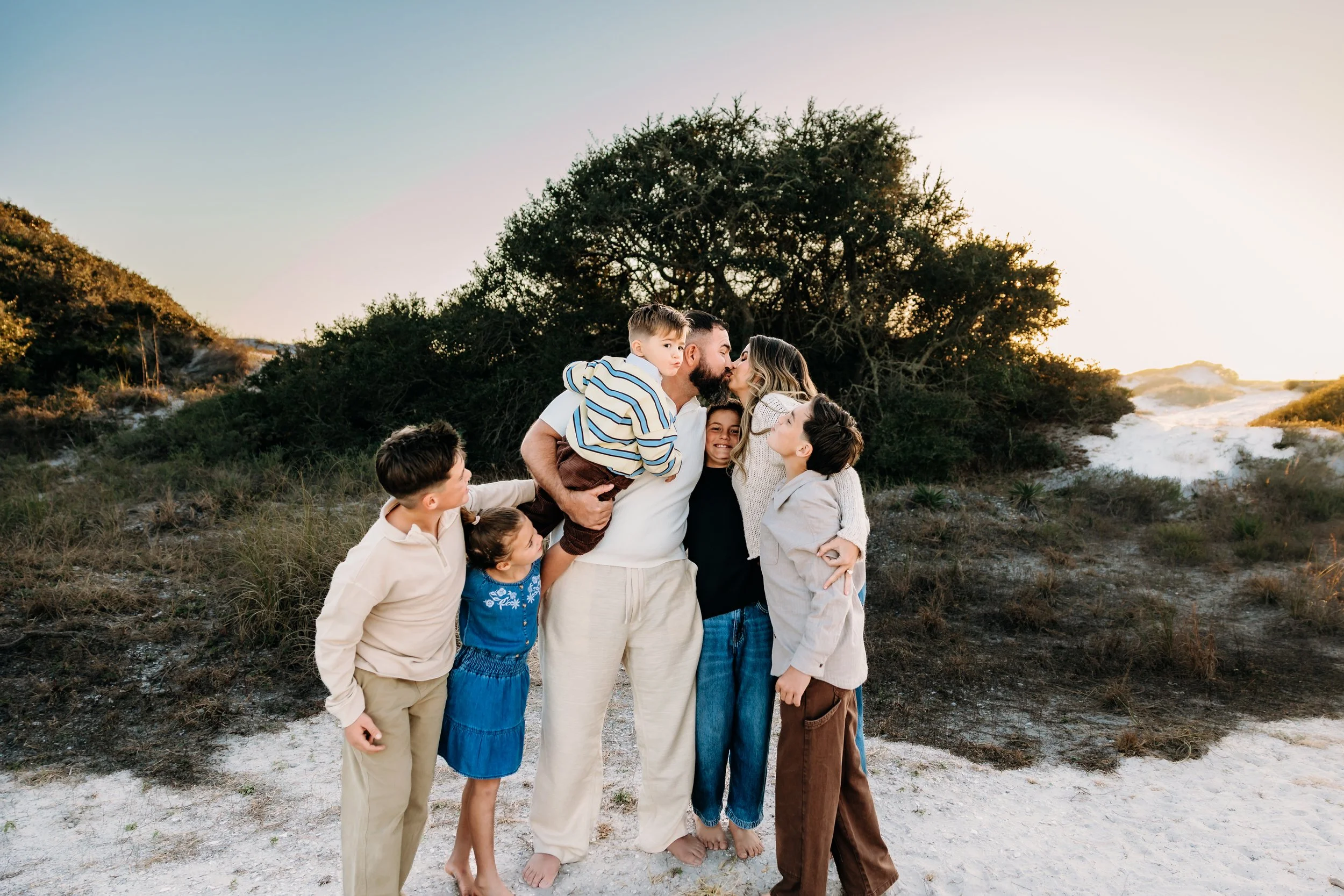 Family of eight people, including adults and children, embracing and kissing outdoors on sandy terrain with bushes and a large tree, during sunset.
