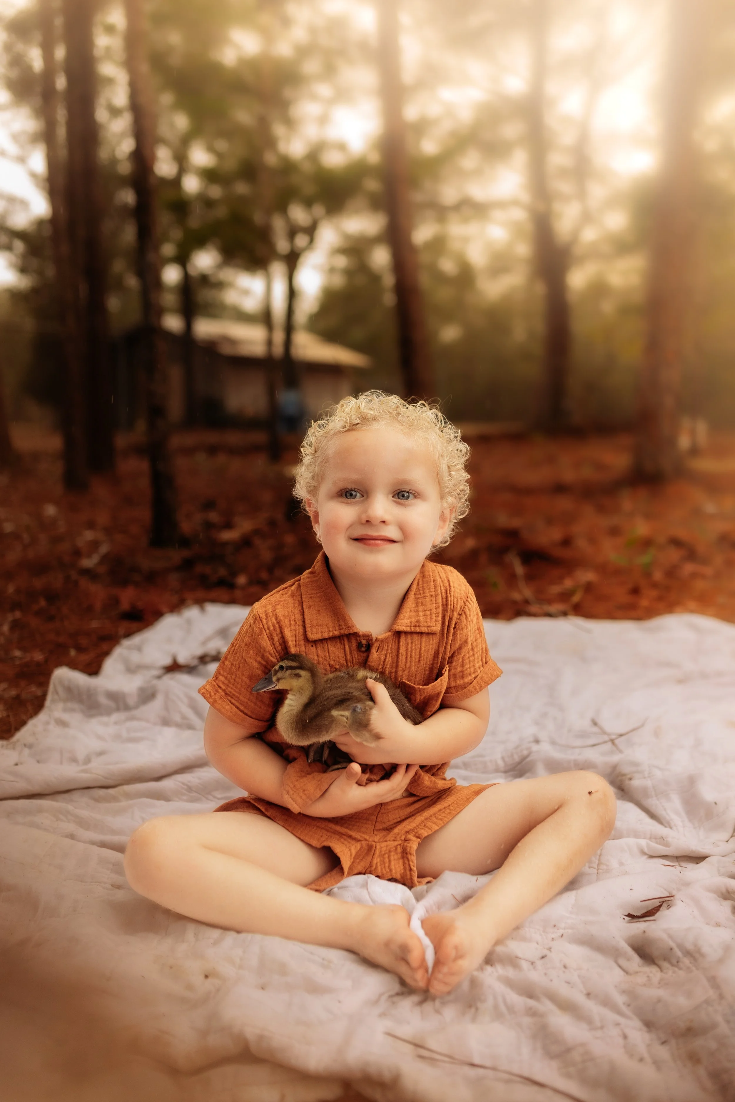 A young boy with curly blonde hair and blue eyes sitting cross-legged on a white blanket outdoors, holding a small duckling. The background features trees with sunlight filtering through the branches.