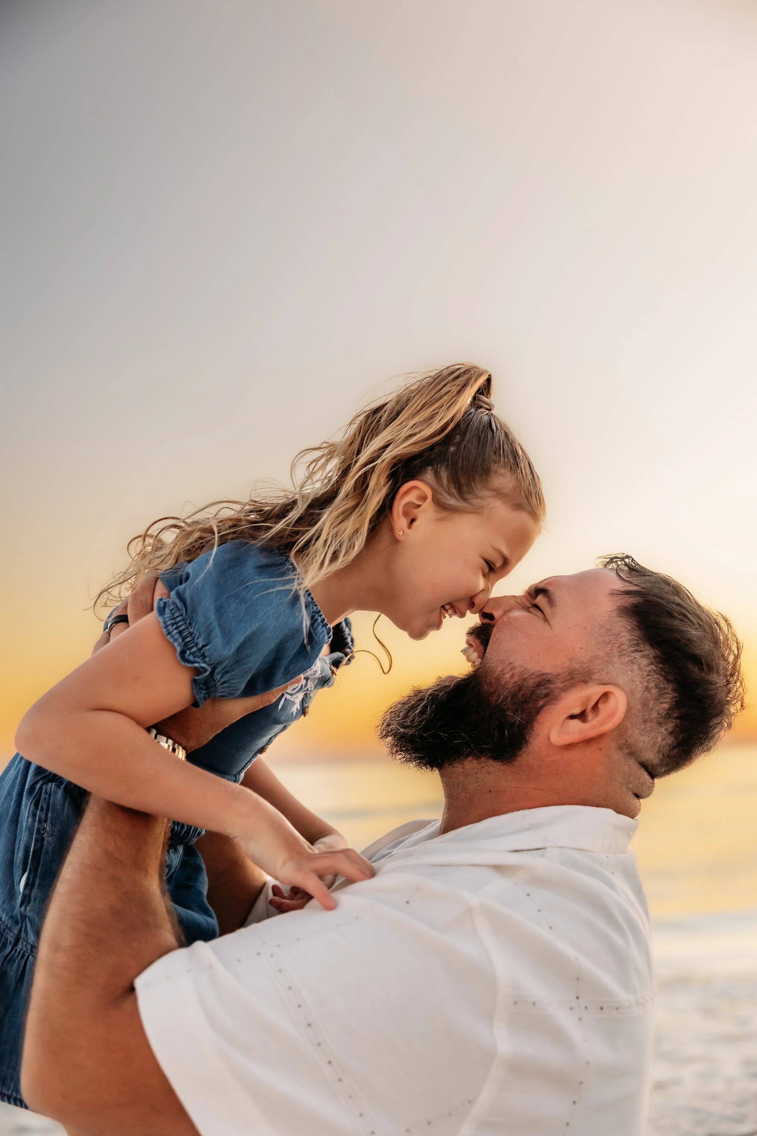 A man lifting a young girl, both smiling and touching foreheads, on a beach at sunset.