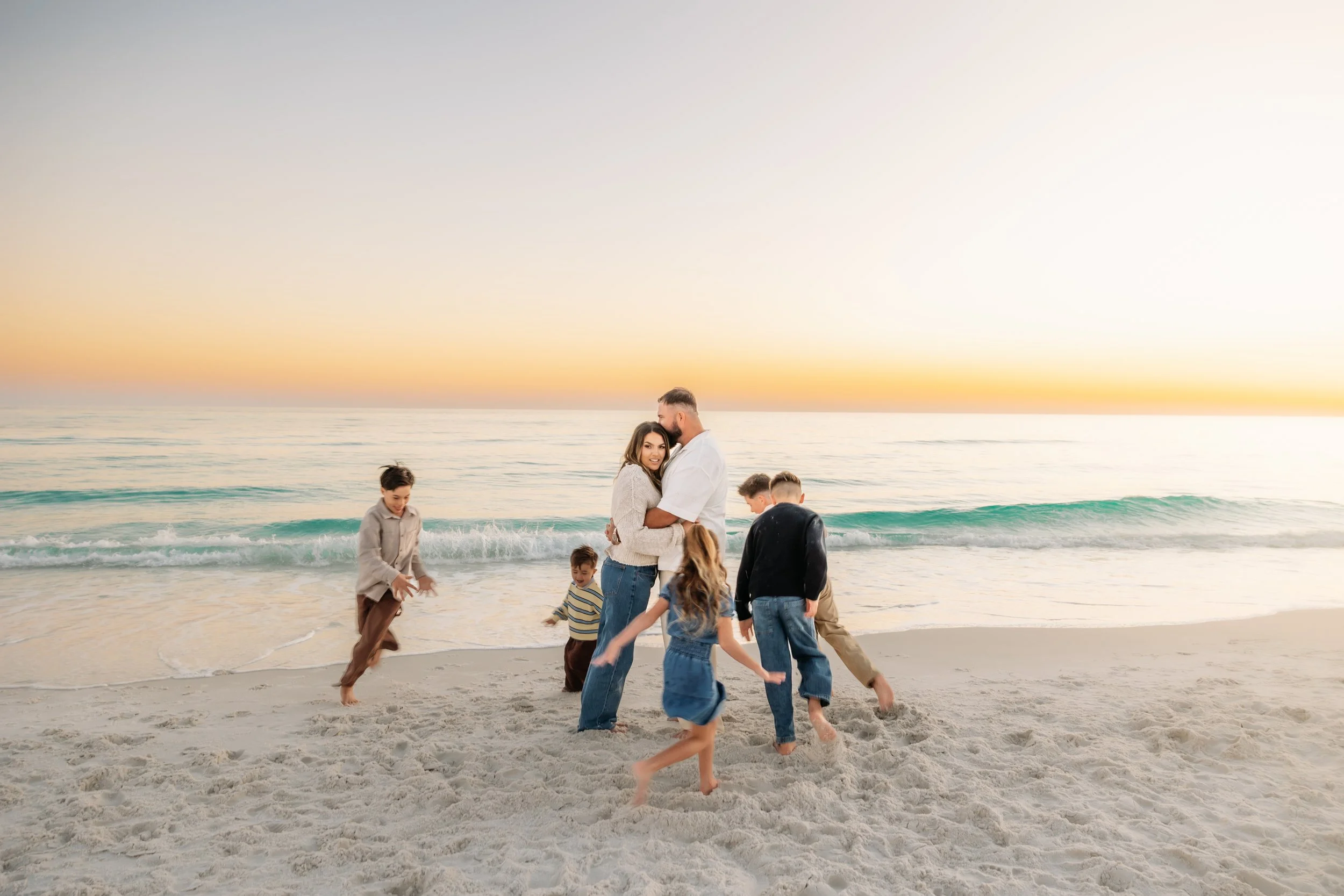 Group of children and two adults playing and walking on the beach during sunset, with ocean waves in the background.