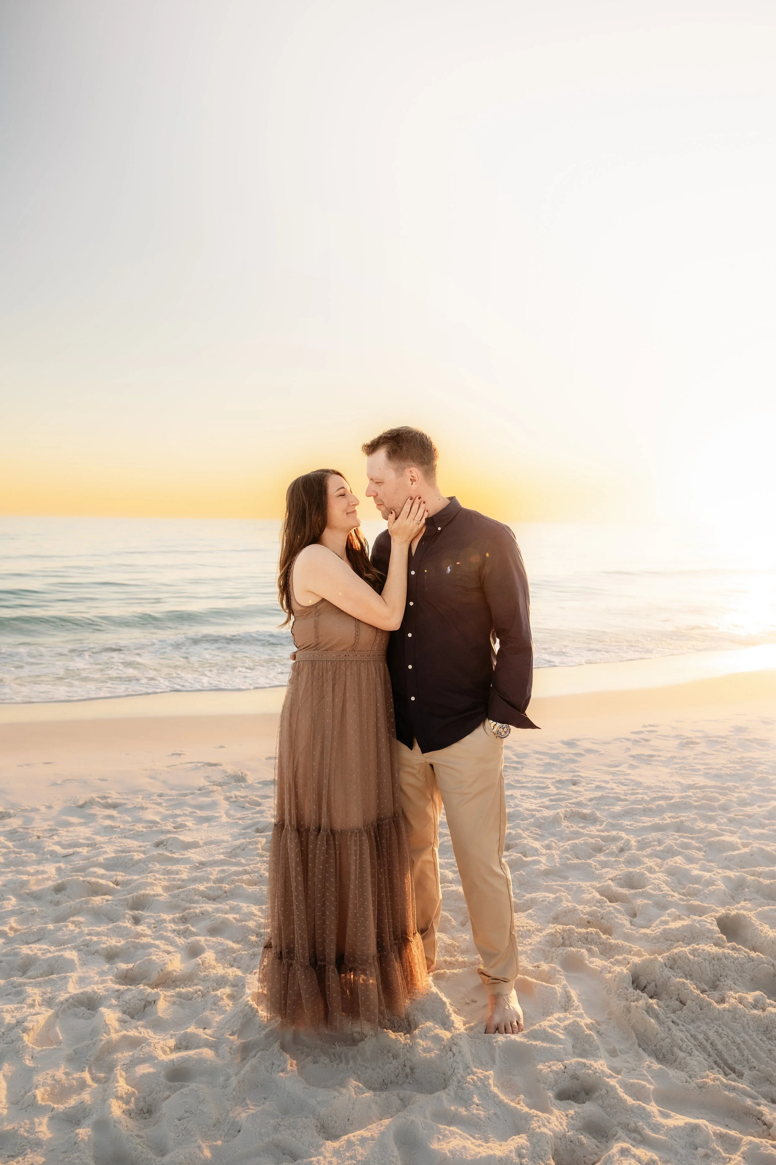 A couple standing close together on a sandy beach at sunset, with the ocean in the background. The woman is touching the man's face and they are smiling at each other.