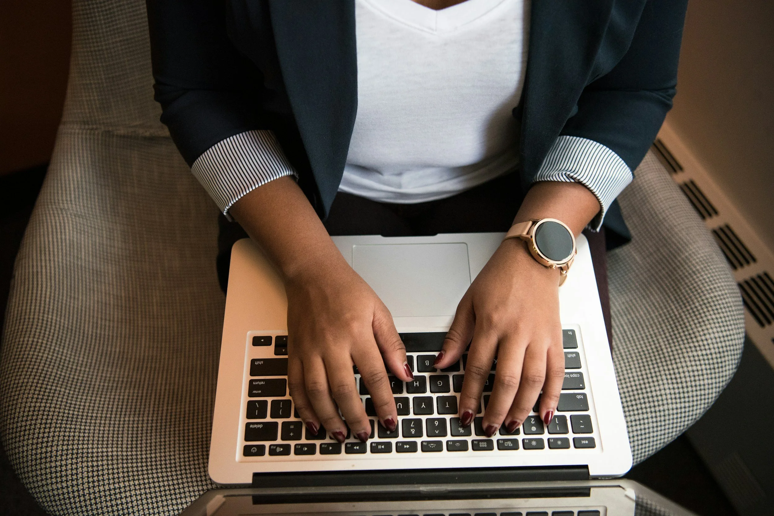 Person working on a silver laptop, sitting on a patterned chair, wearing a dark blazer with striped cuffs, a white shirt, and a smartwatch.