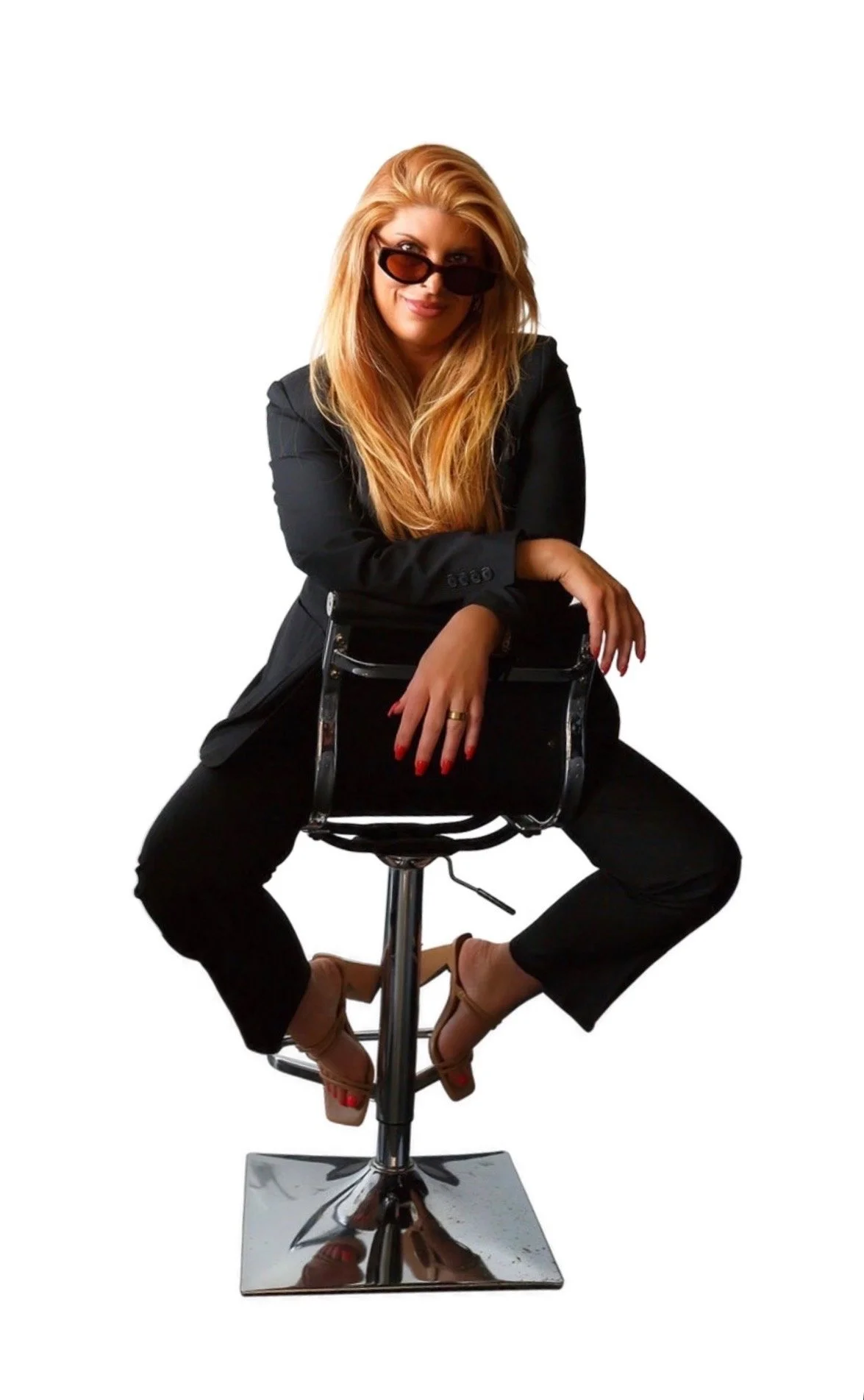 A woman with long red hair wearing sunglasses, a black blazer, and black pants, is sitting on a black and chrome adjustable stool, posing with her right arm resting on the back of the stool and her left hand on top of it, against a plain white background.