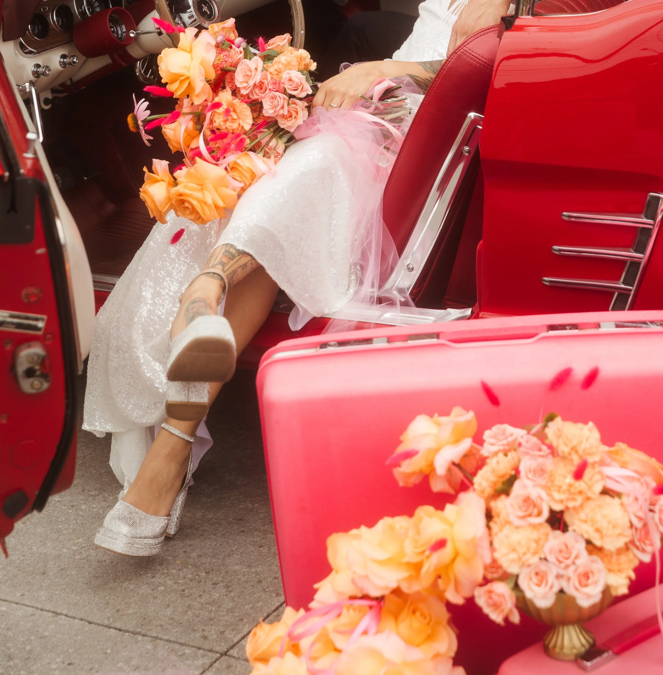 A bride lying in a vintage red car, holding a bouquet of peach and pink flowers, with the car's interior and pink floral arrangements visible.