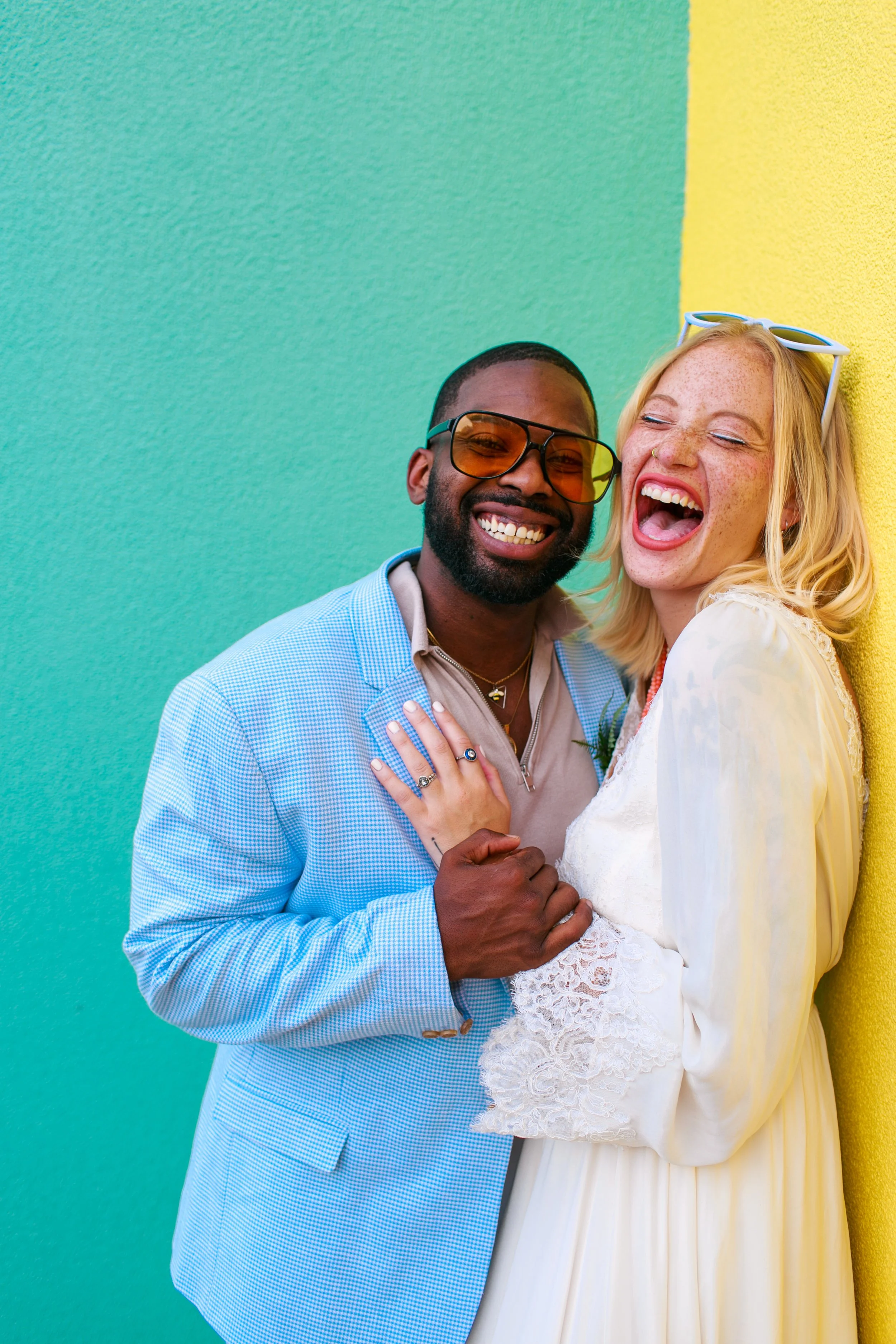 Joyful couple laughing, standing against colorful green and yellow wall, the woman with blonde hair and sunglasses on head, the man with dark skin and beard, both dressed stylishly.