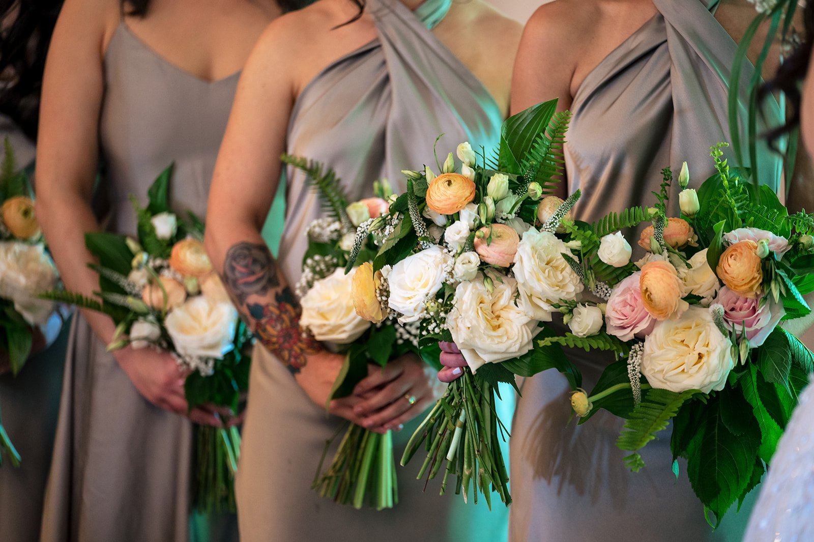 Group of women in matching taupe dresses holding bouquets of white and peach flowers with green leaves, at a wedding or formal event.