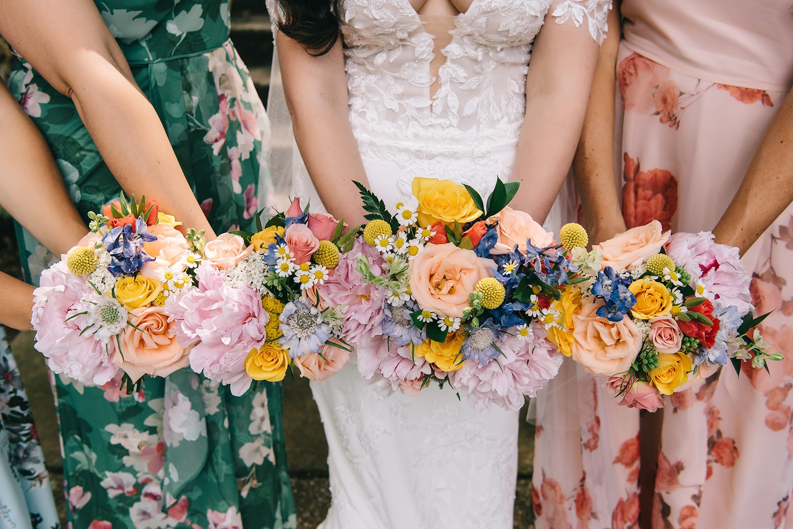 Close-up of women holding a colorful floral bouquet, with a bride in a white lace dress in the center, surrounded by women in floral dresses.