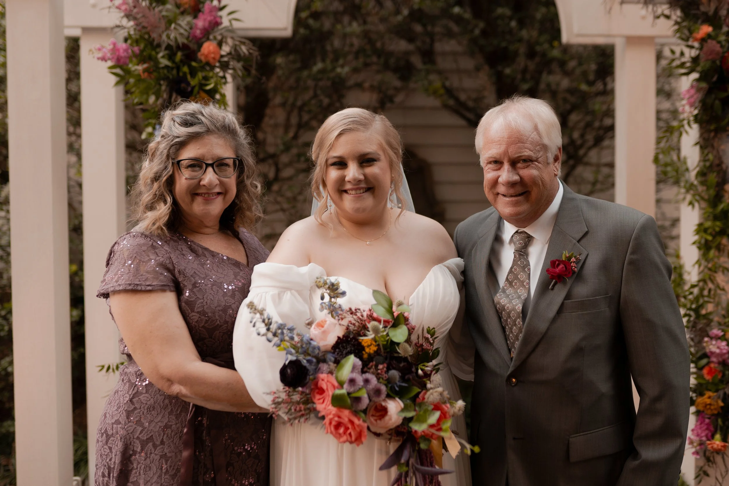 A bride standing between her mother and father, smiling, holding a bouquet of flowers, with floral decorations in the background at a wedding.