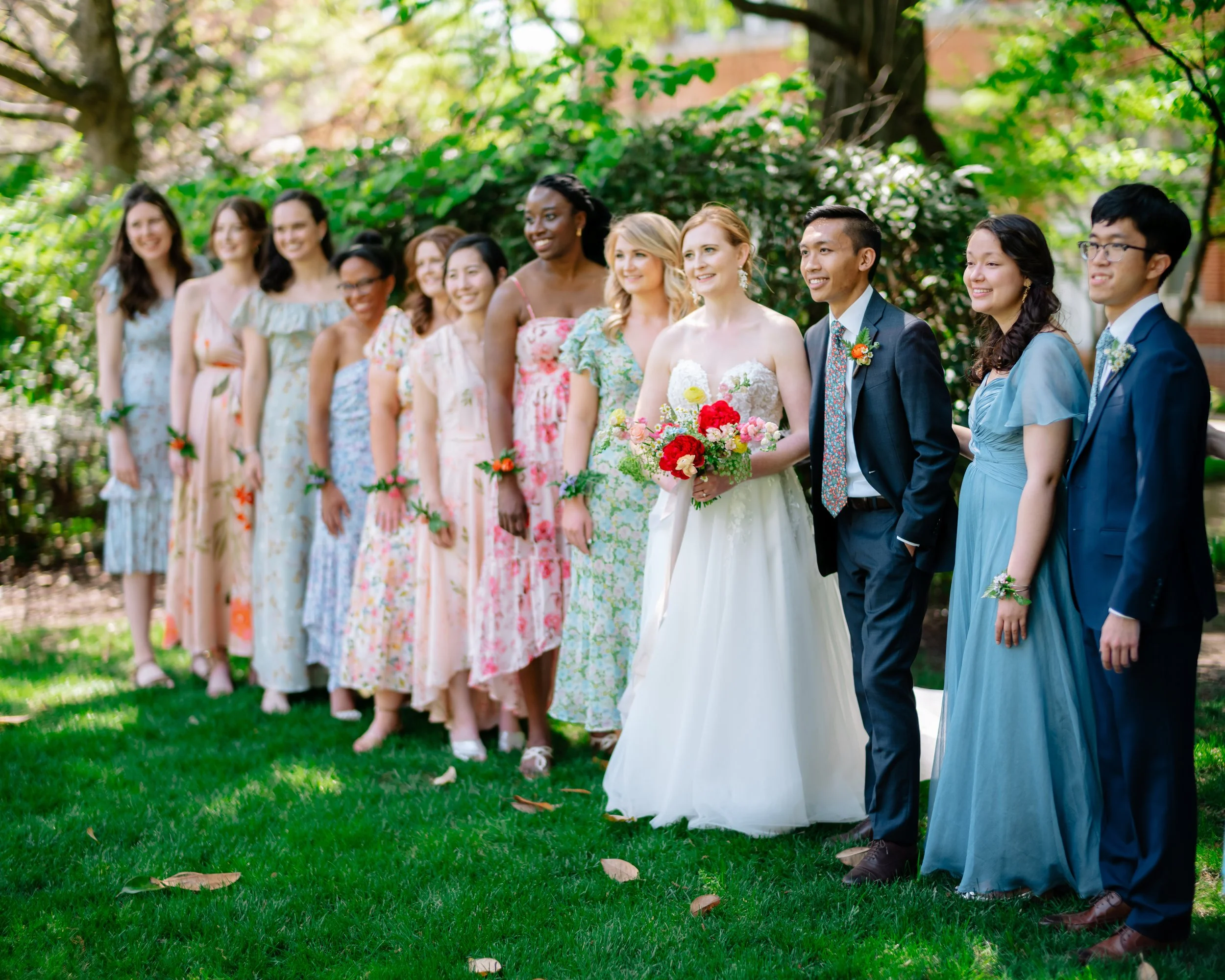 Group of people at a wedding ceremony outdoors, including a bride in a white wedding gown holding a bouquet of flowers, surrounded by bridesmaids and groomsmen in colorful dresses and suits, with green trees and grass in the background.