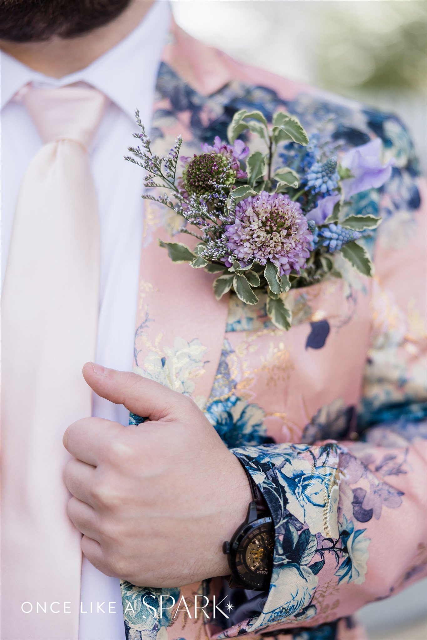 A man wearing a floral pink blazer, white shirt, and pink tie, holding a boutonniere made of purple daisies, lavender, and other flowers. He is also wearing a black watch.