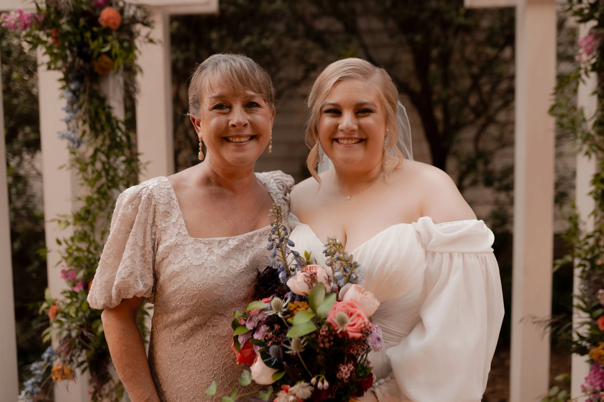 A bride and an older woman, possibly her mother, standing together outdoors on a wedding day. The bride is holding a bouquet of flowers and wearing an off-the-shoulder wedding dress, while the older woman is dressed in a lace dress. Both are smiling